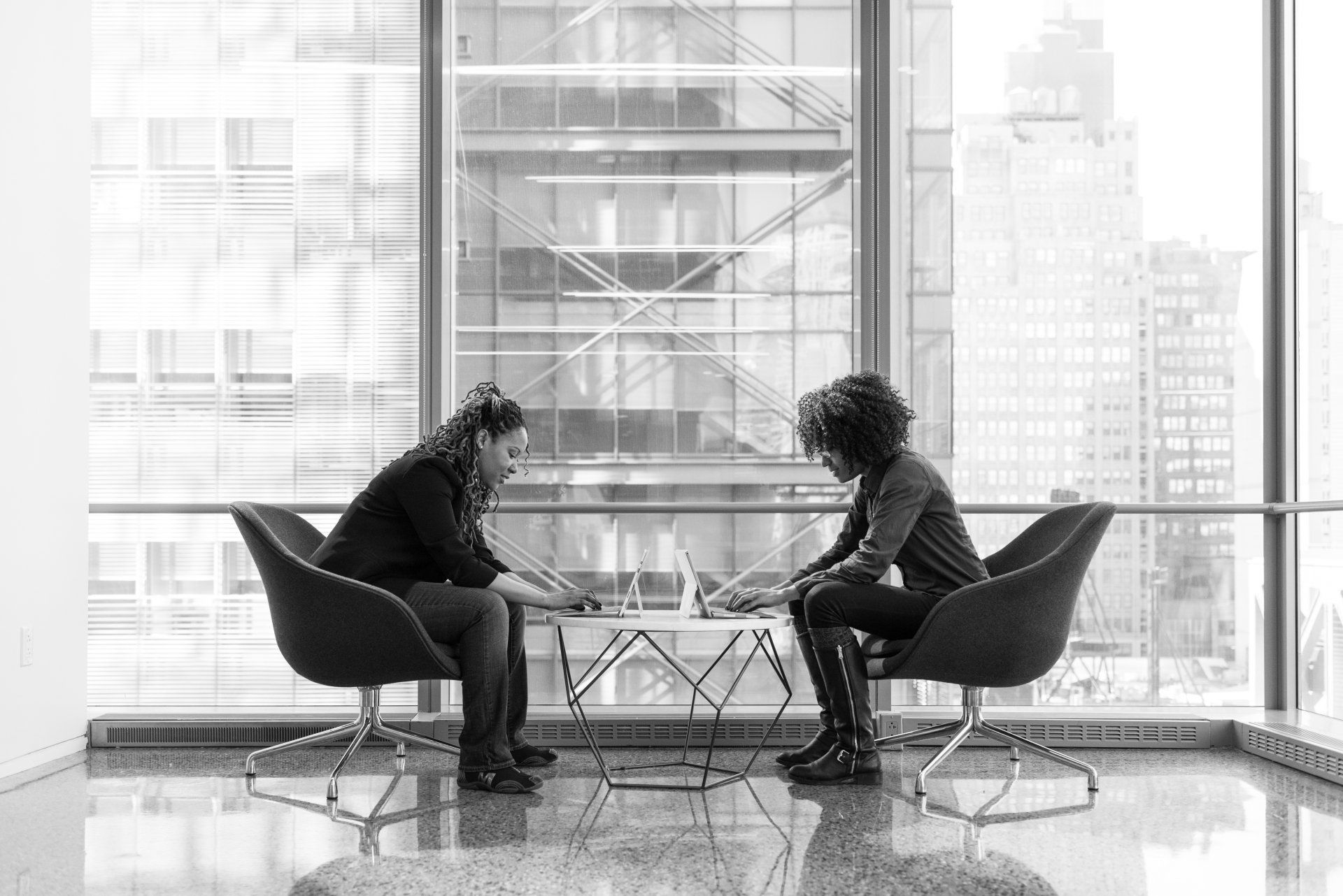 Two black females sitting across from one another at a short round table. Both are working on laptops.