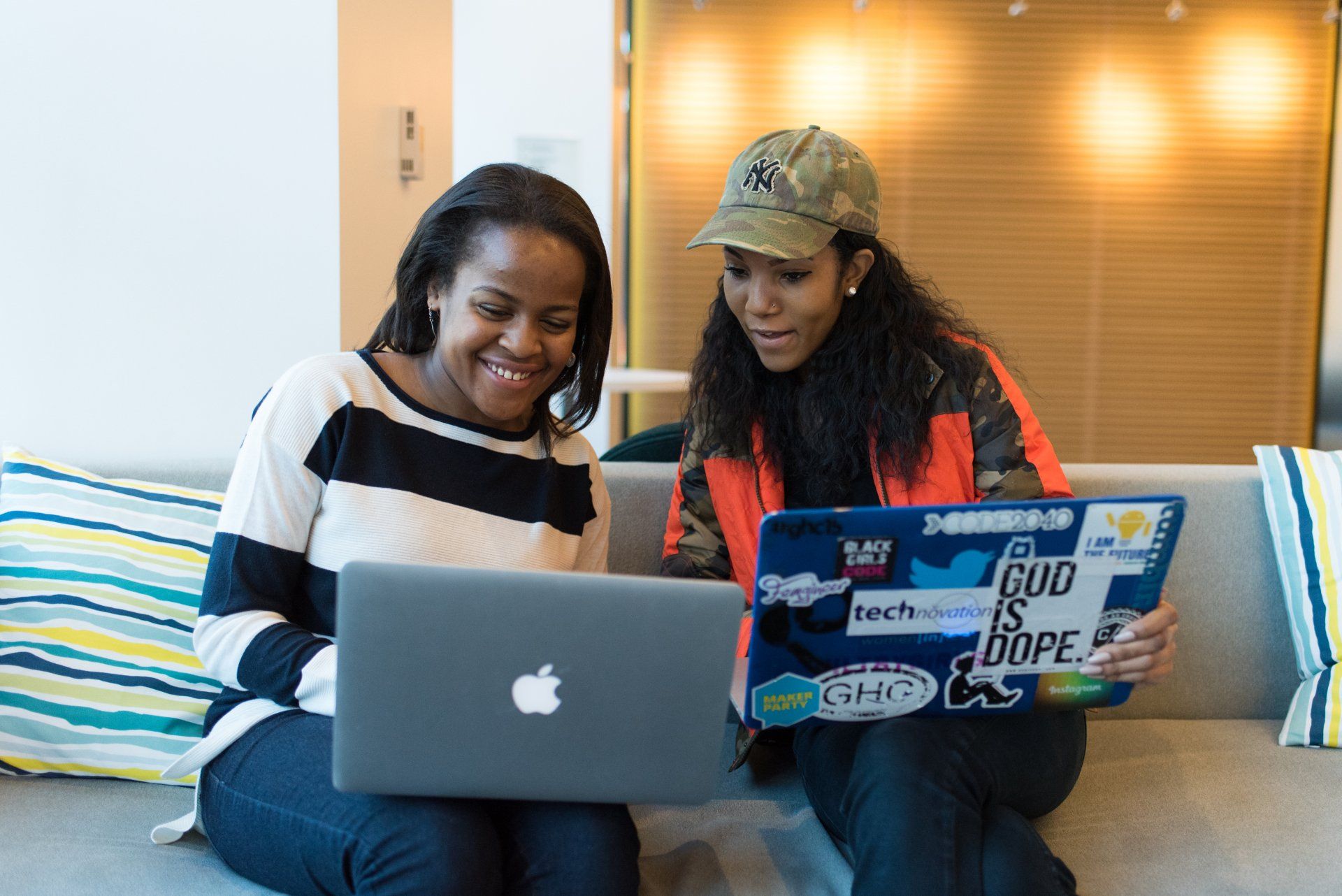 Two Black women sitting on a couch with open computers talking to each other.