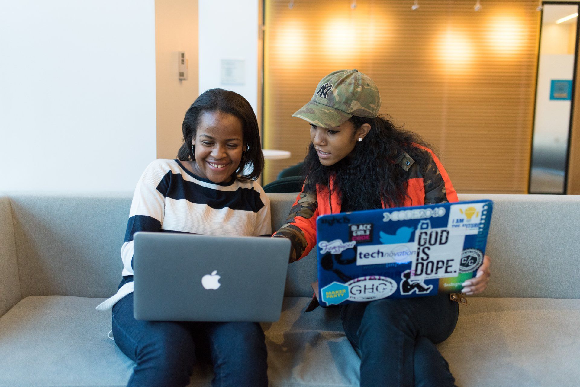 Two women are sitting on a couch looking at laptops.