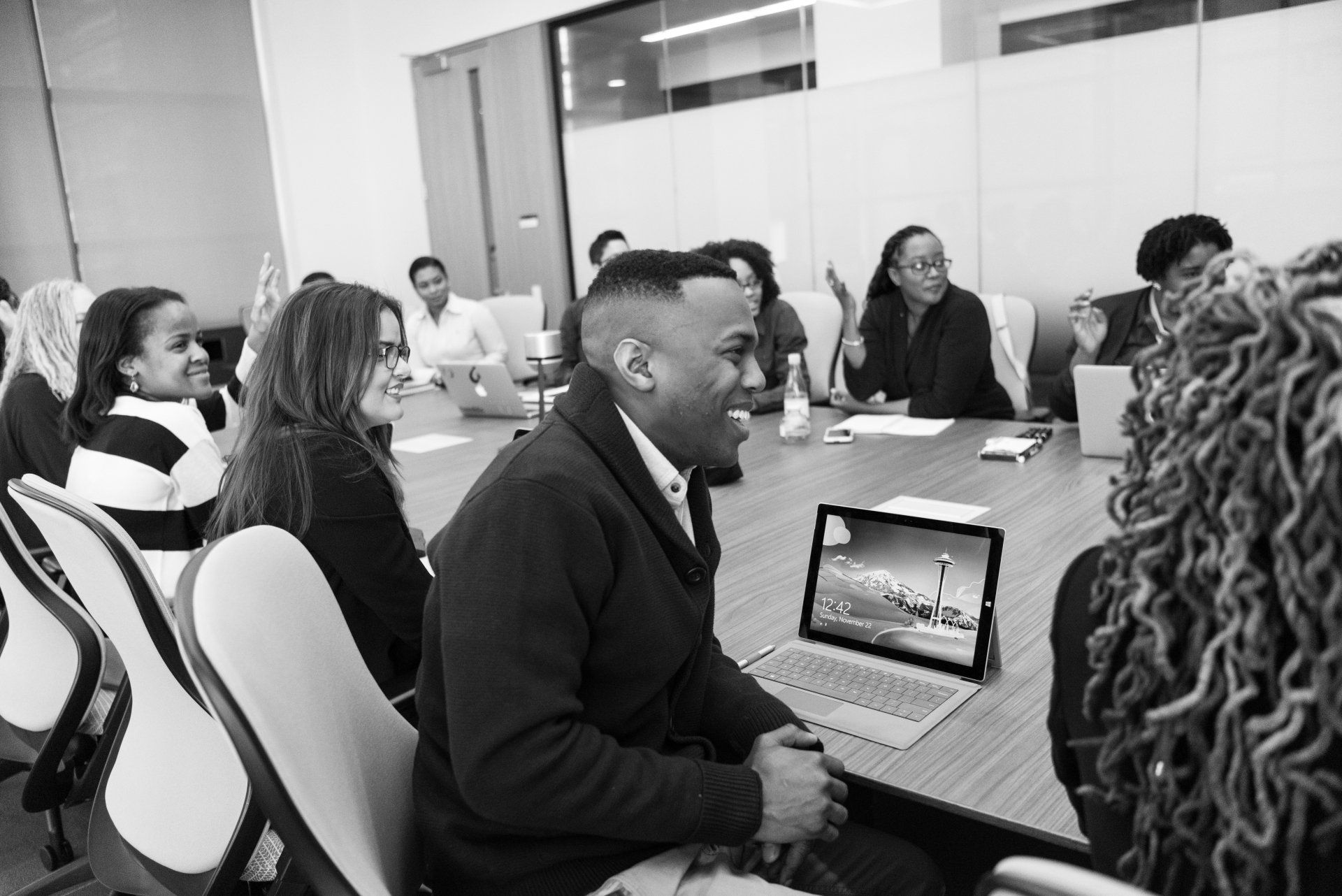 Black and white image of race, gender, age group of at least 10 people sitting around a conference table looking at/engaging with something or someone beyond the scope of the image