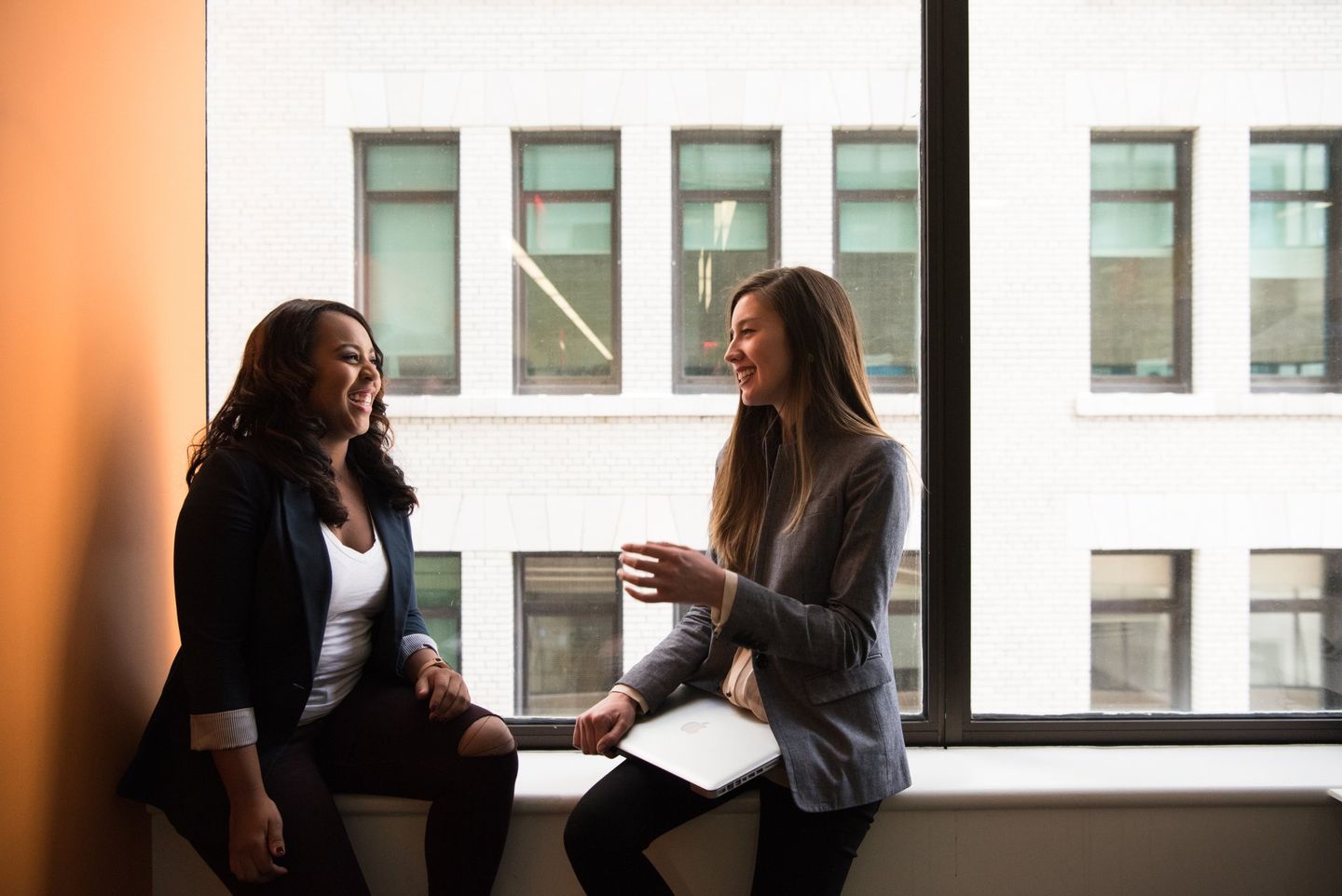 Two women are sitting on a window sill talking to each other.