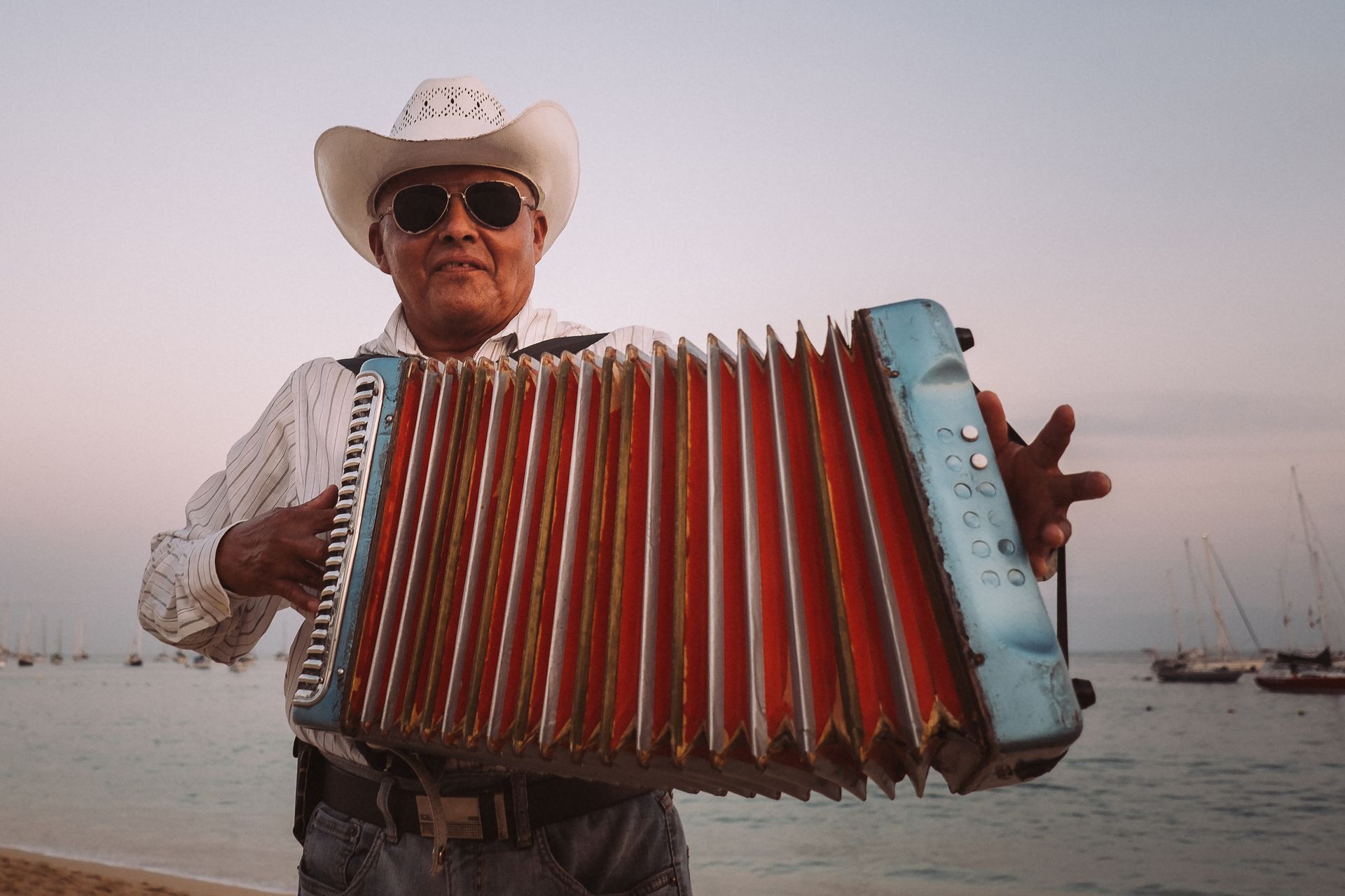 Un homme portant un chapeau de cow-boy joue de l'accordéon bleu et orange sur une plage.