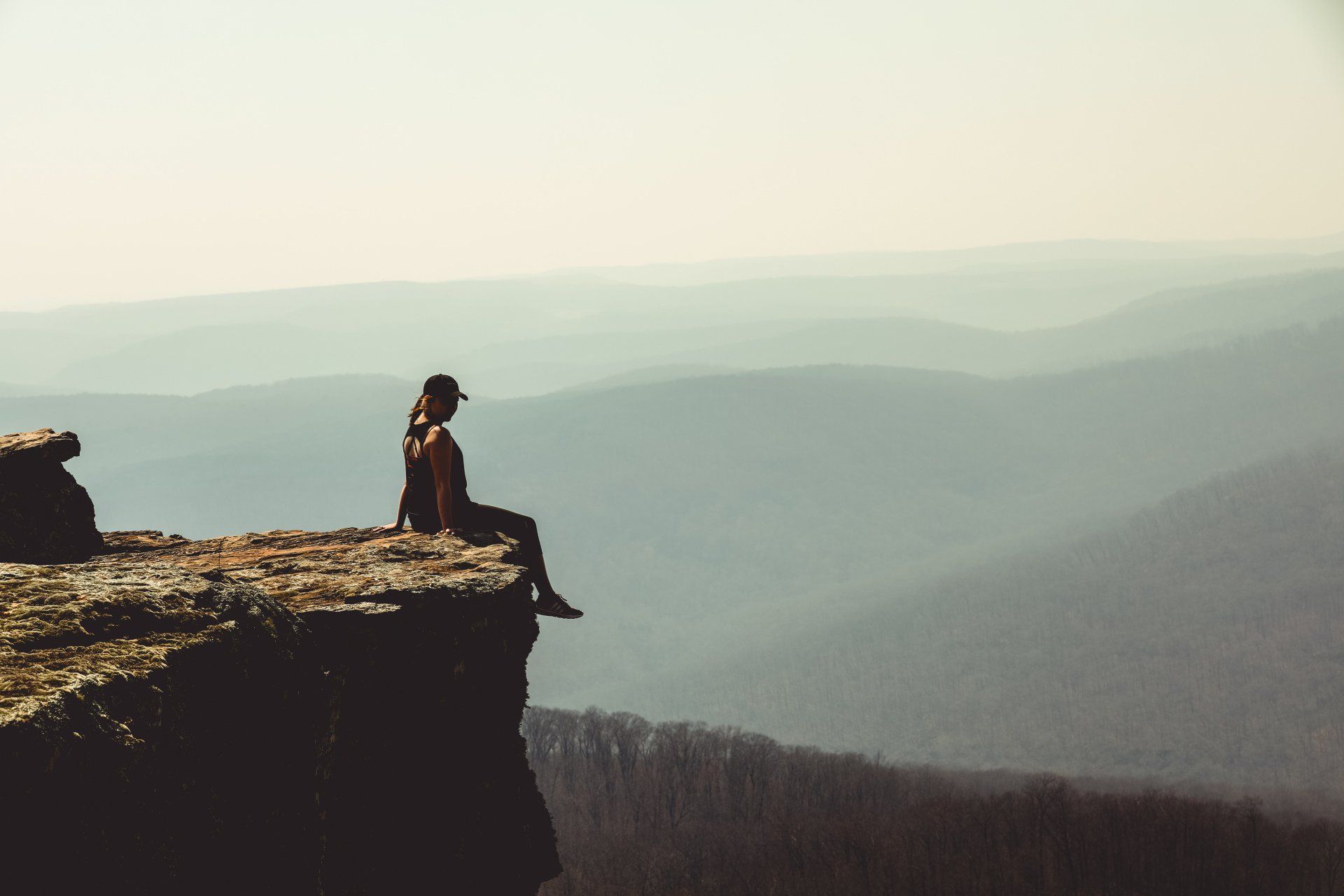 A person is sitting on the edge of a cliff overlooking a mountain range.