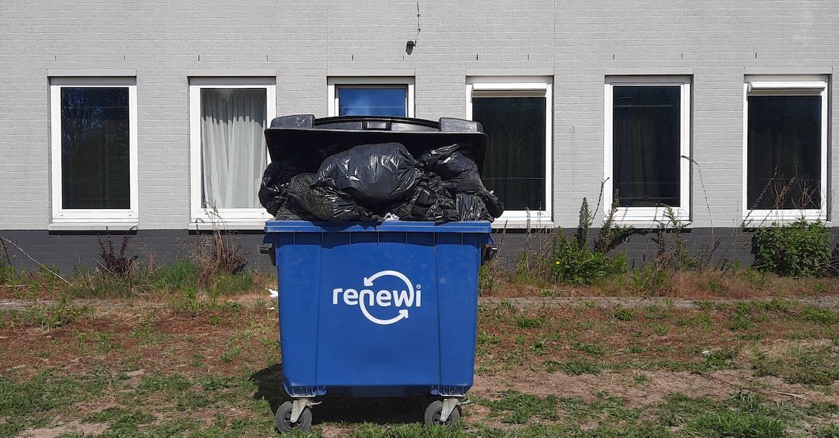 A blue trash can is filled with trash bags in front of a building.