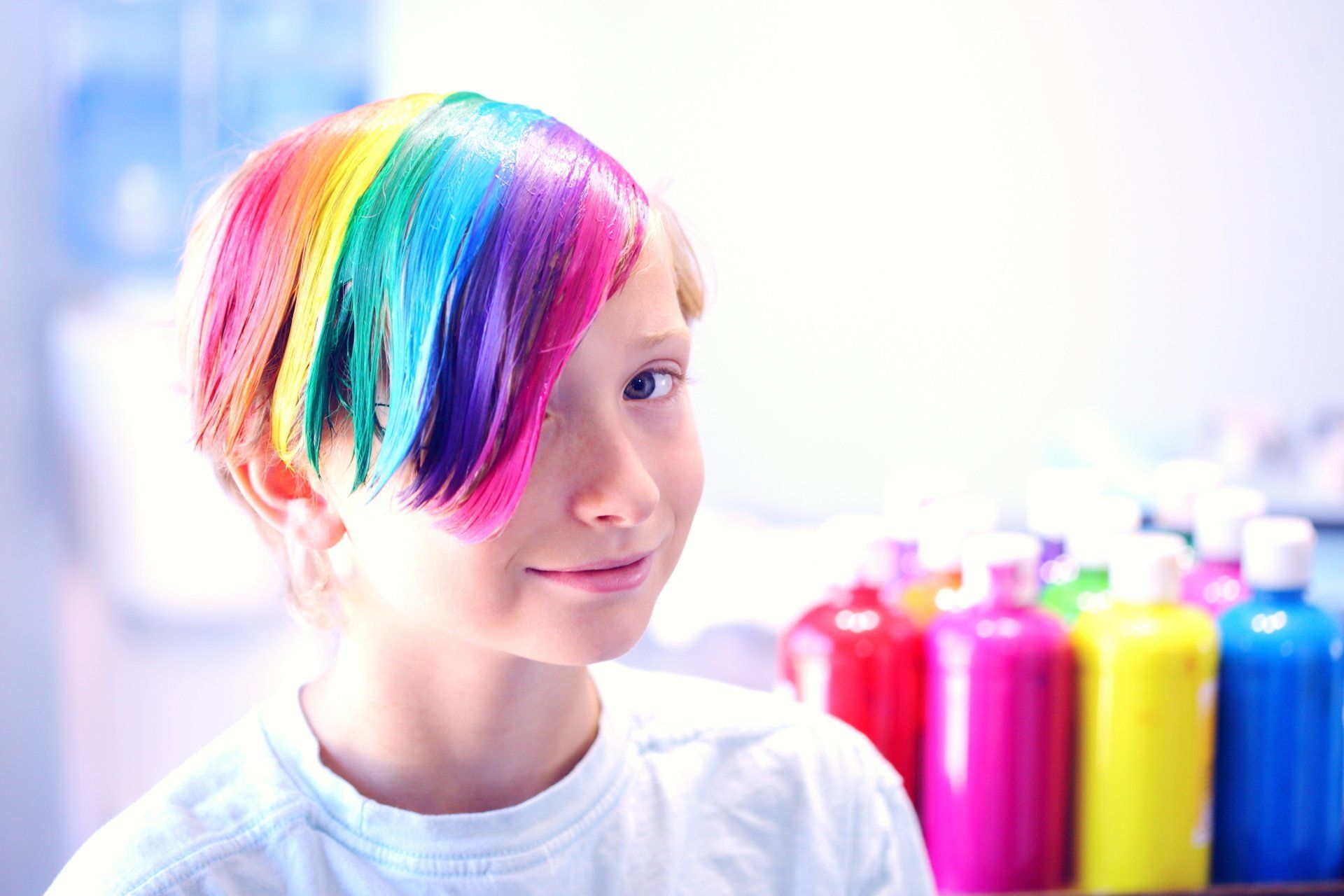 A young girl with rainbow hair is sitting in front of bottles of paint.