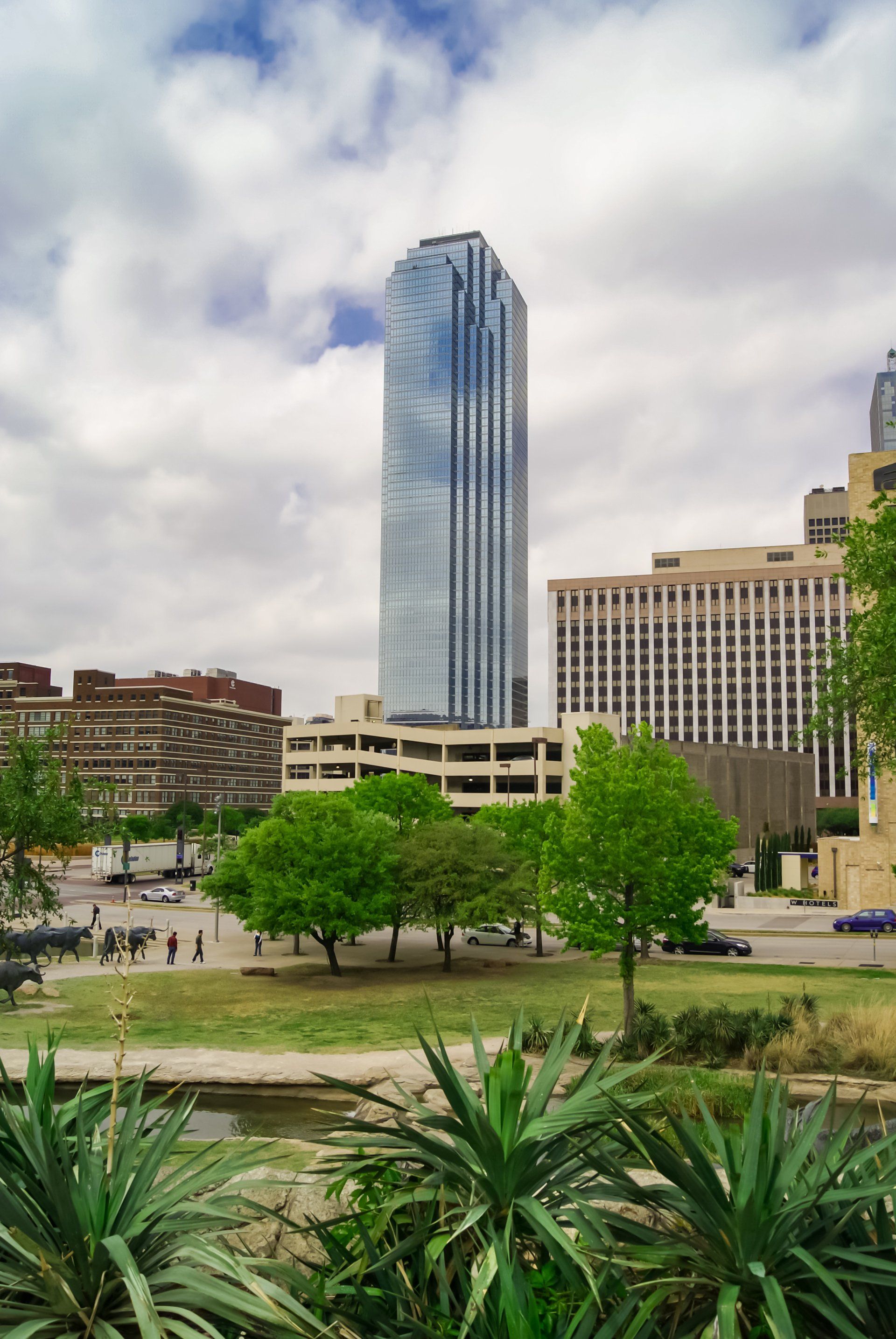 A modern skyscraper dominates a city skyline with trees in the foreground and a cloudy sky above.