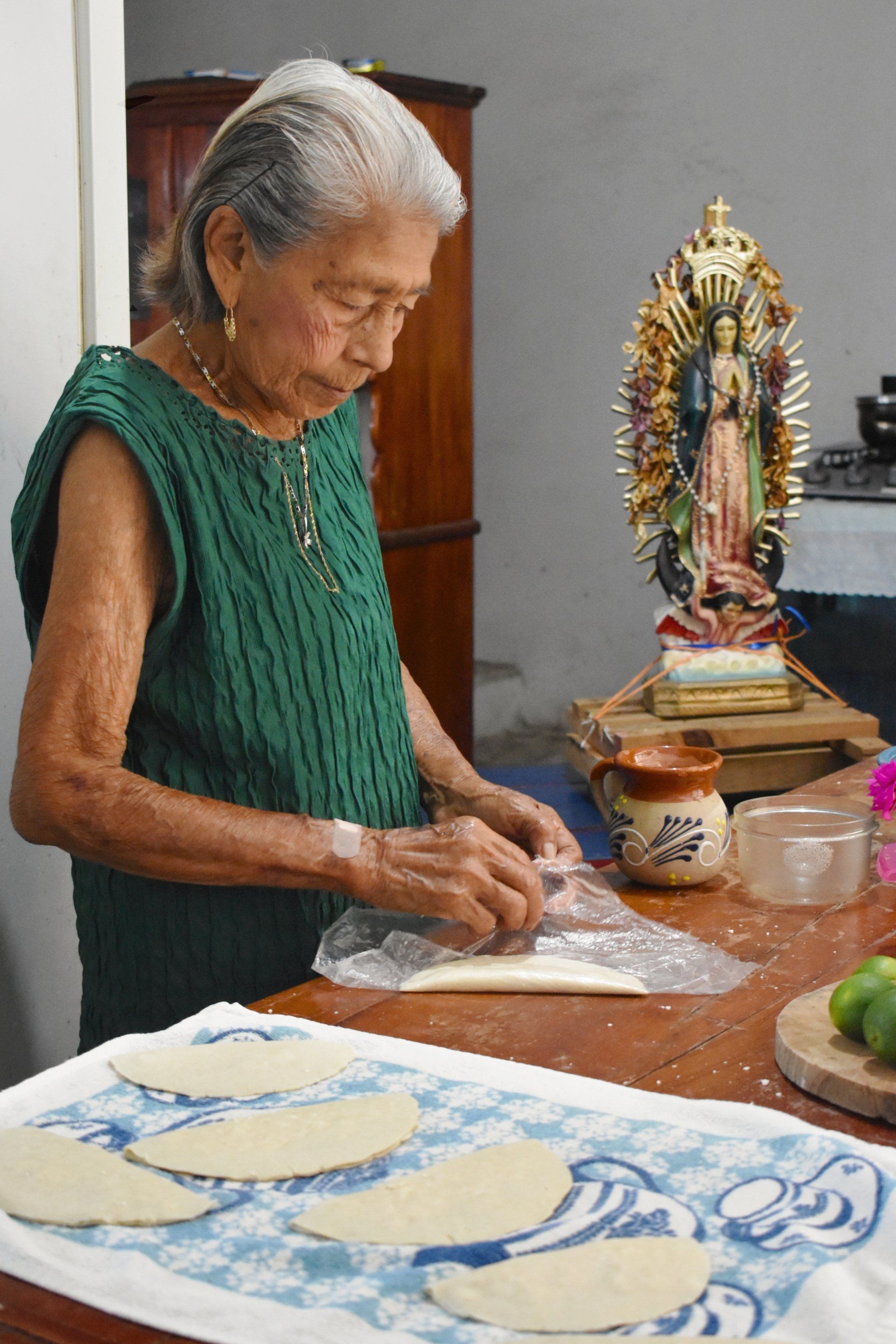 An elderly woman is making tortillas on a table in front of a statue.