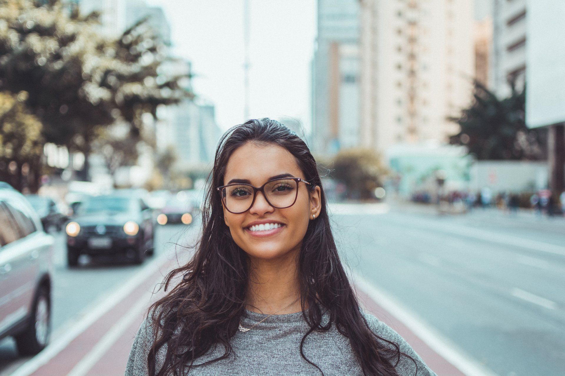 A woman wearing glasses is smiling in front of a city street.