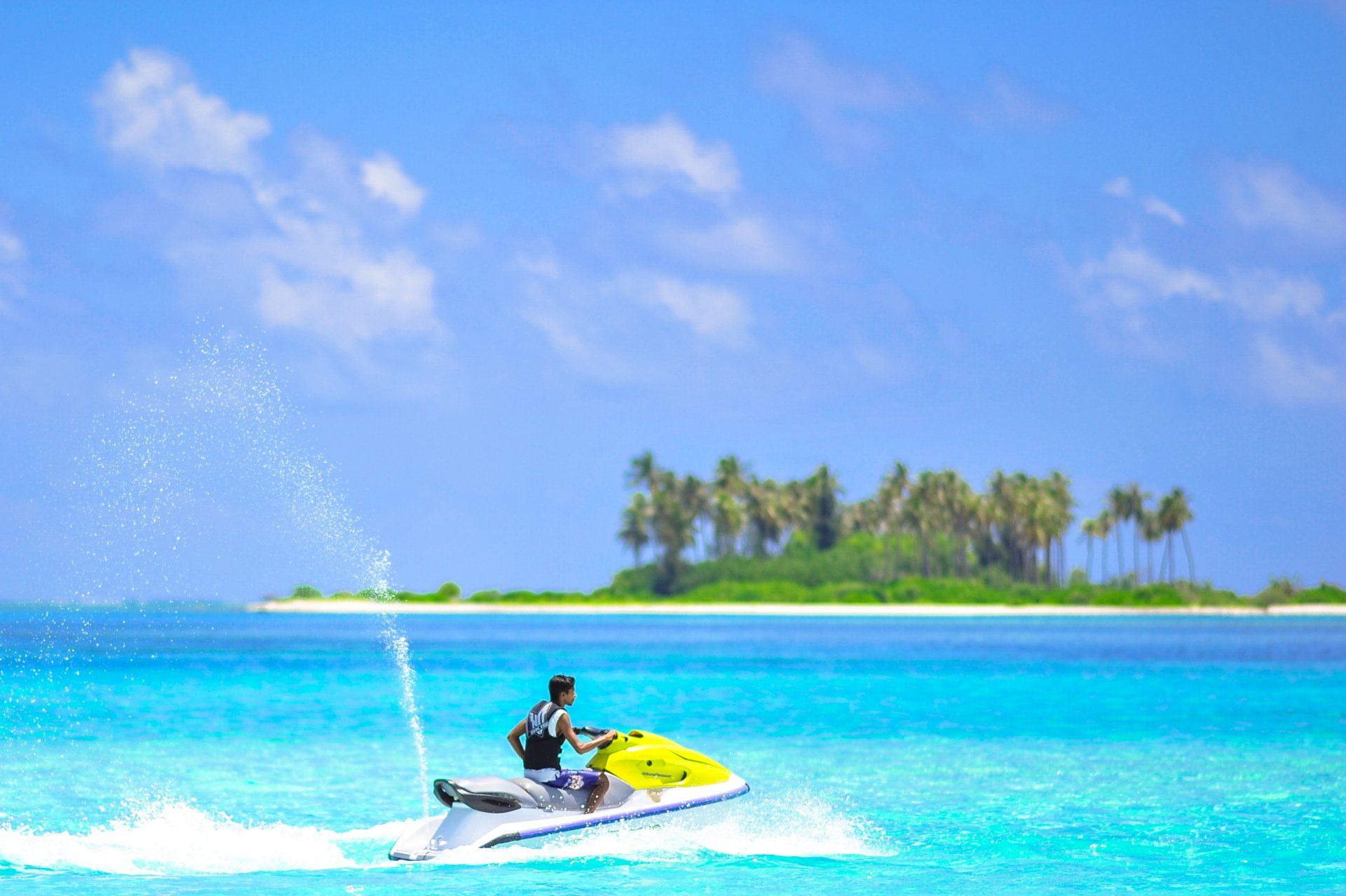 Man on jet ski in turquoise water, island with palm trees in background, sunny day.