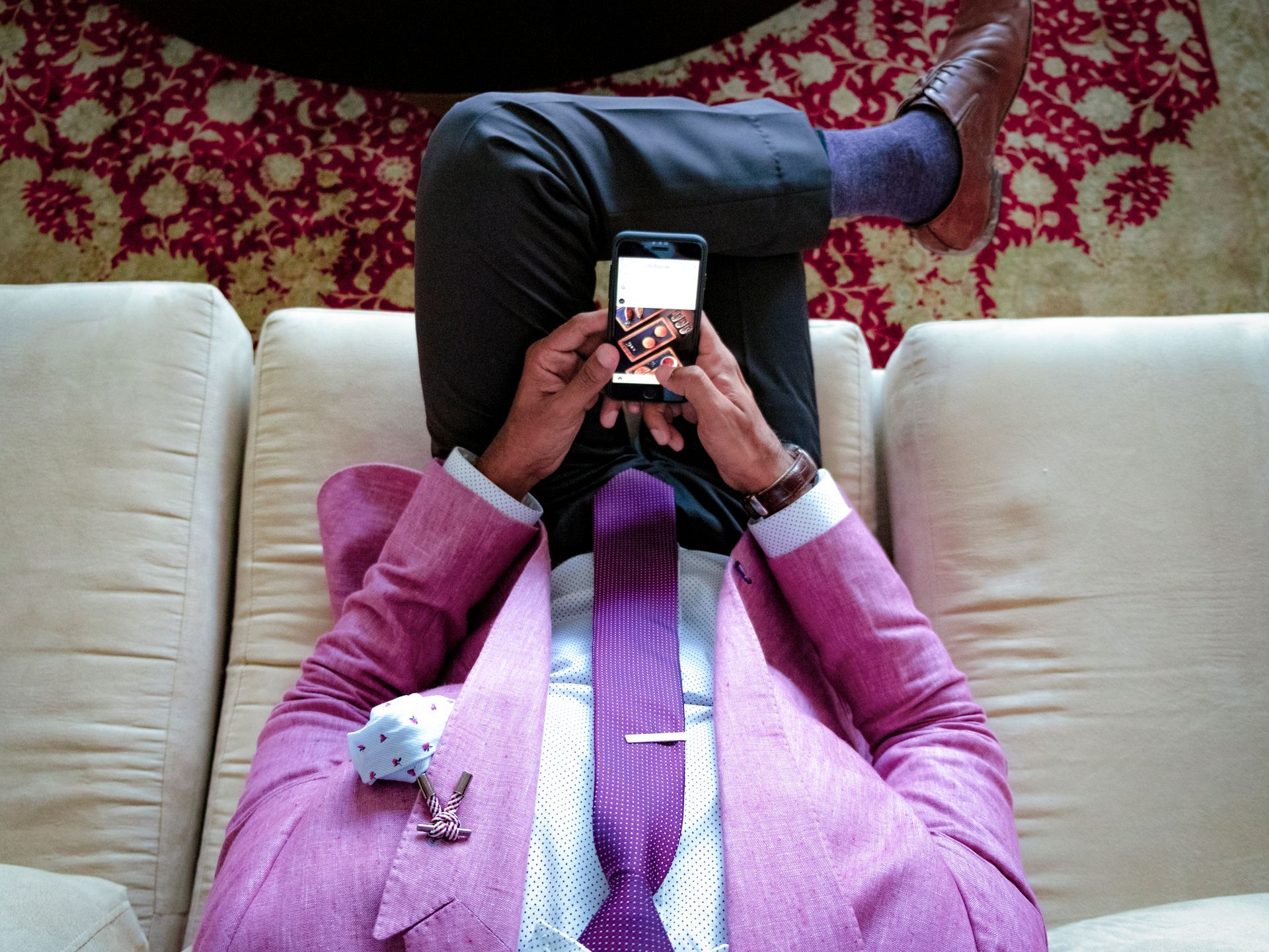 Man in pink suit, holding phone, reclining on a white couch. Purple tie, socks, and loafers.