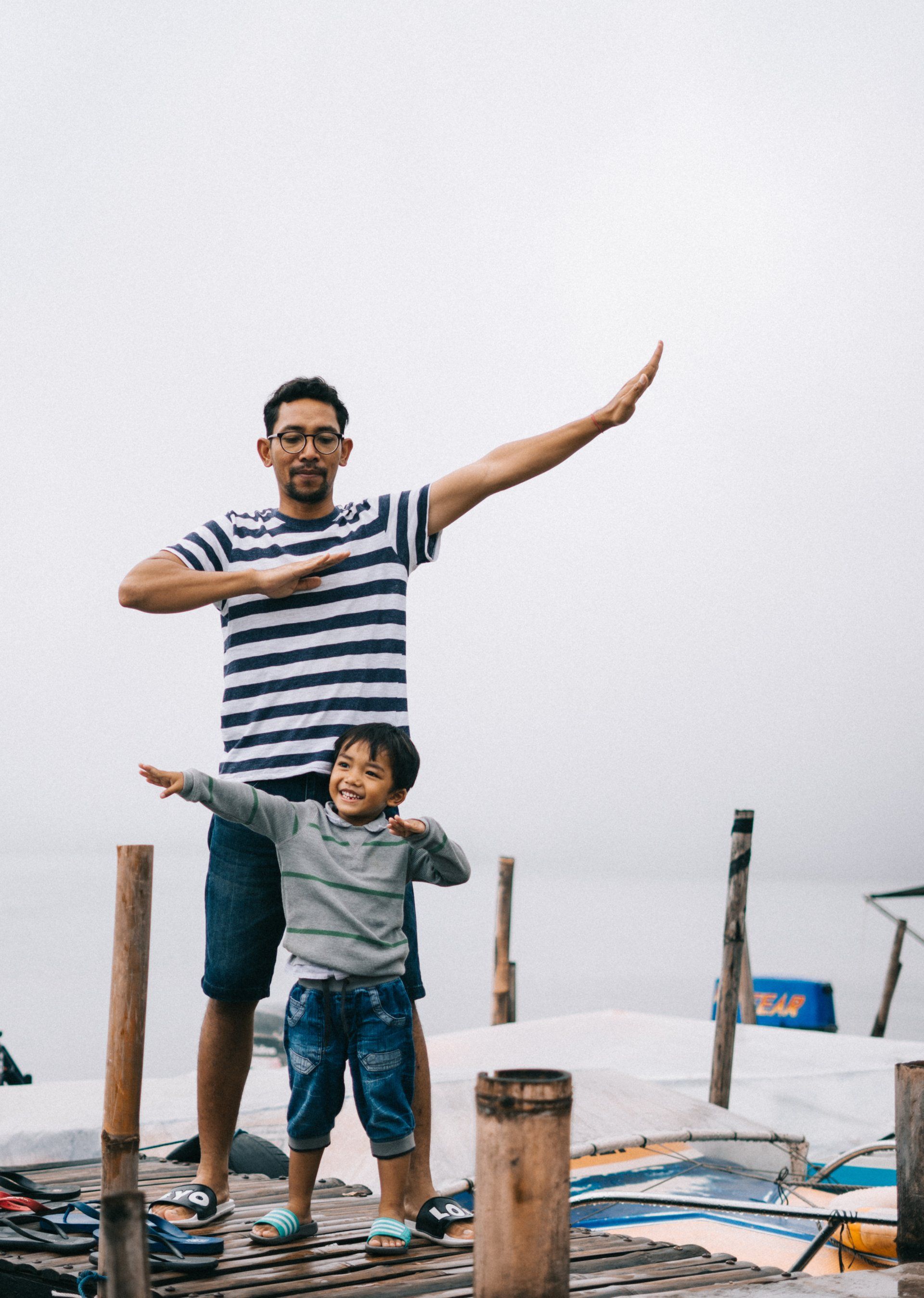 A man and a child are standing on a dock with their arms outstretched.