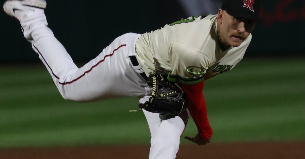 A baseball pitcher in white pants and a light-colored jersey throws a ball on a green field at night.