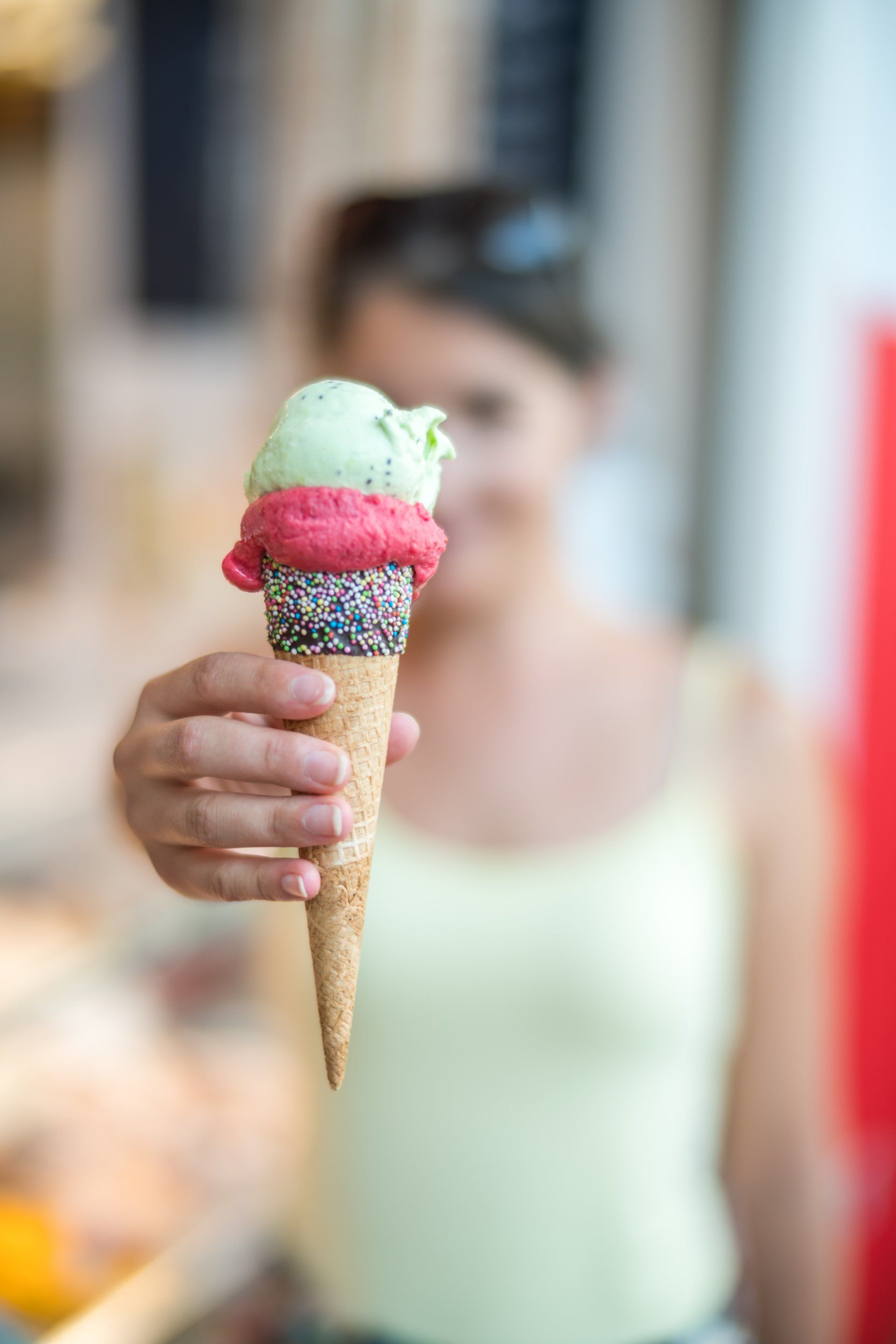Woman holds out a three-scoop ice cream cone with pistachio, raspberry, and chocolate sprinkles.
