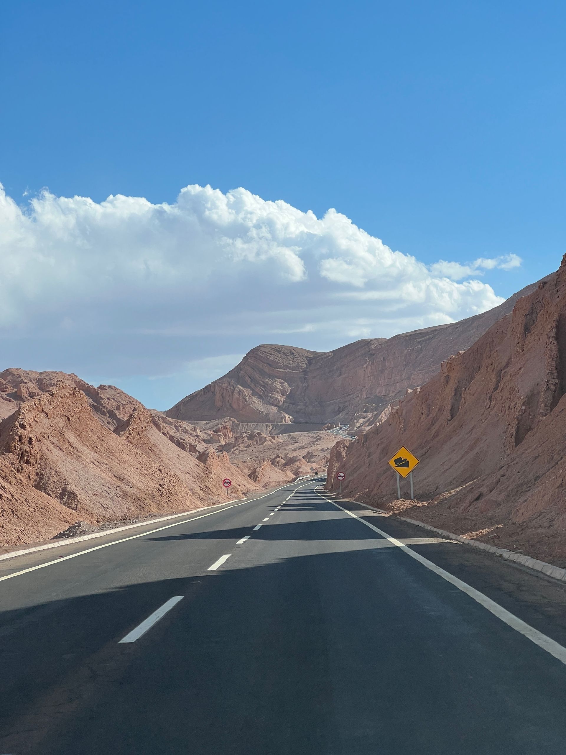 Carretera asfaltada atravesando montañas rojizas bajo un cielo azul con nubes blancas. Señal de tráfico amarilla.