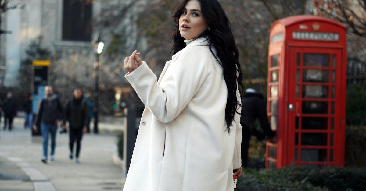 A woman in a white coat is standing in front of a red telephone booth.