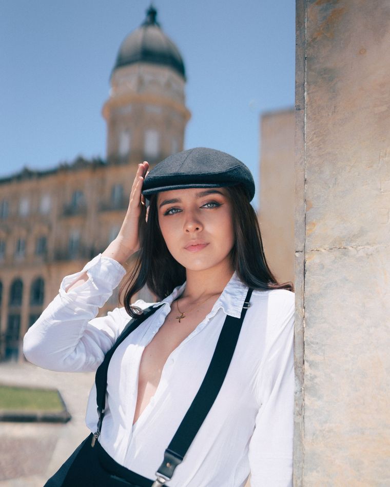 A woman wearing a hat and suspenders is leaning against a wall
