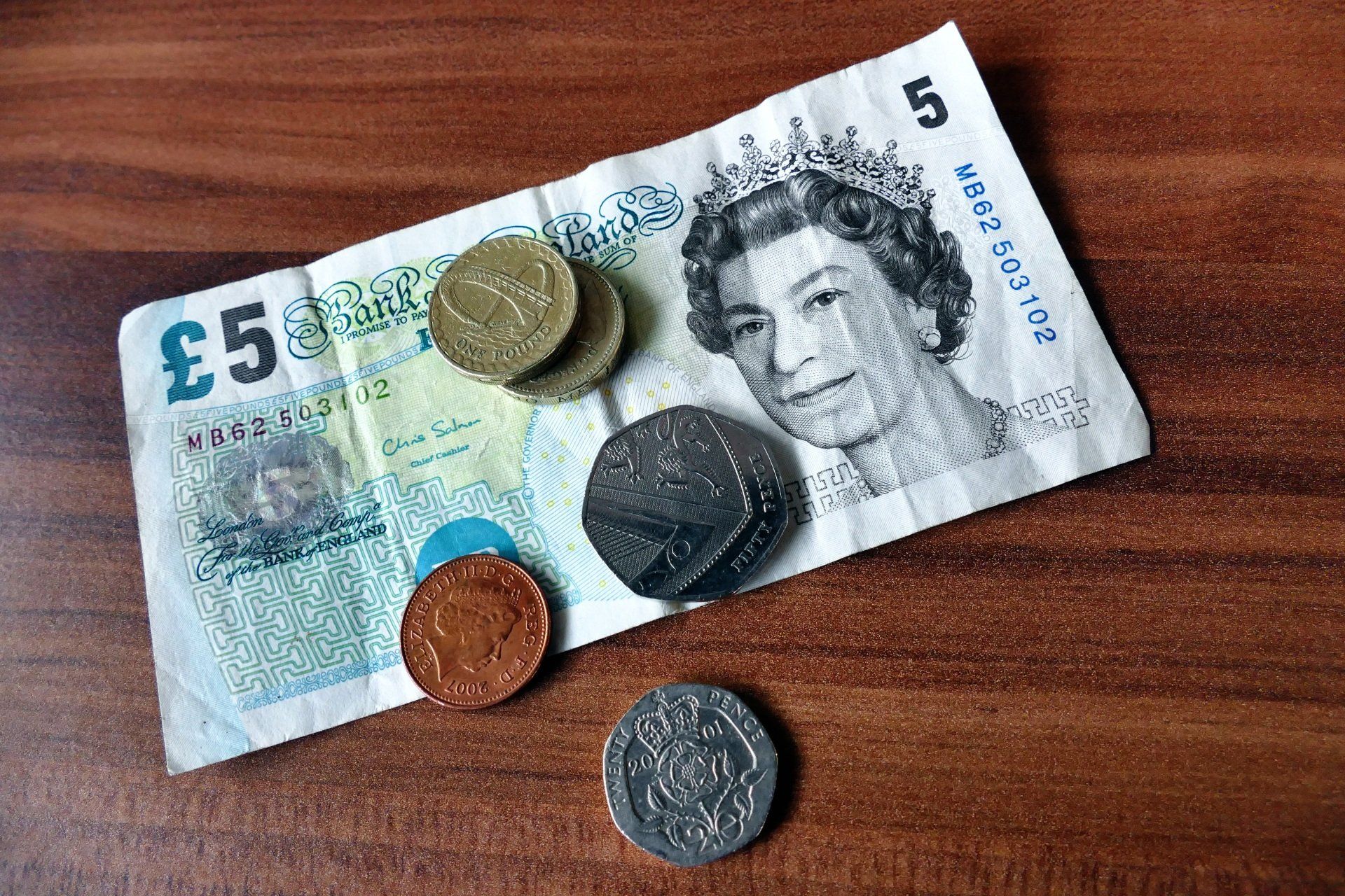 A picture of a £5 note, with 2 x £1 coins stacked on top of each other, 1p coin a 50p coin and a 20p coin on a desk
