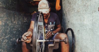 Een persoon repareert een fietswiel in een werkplaats. Hij draagt een pet en werkt met gereedschap, waarschijnlijk voor reparaties.