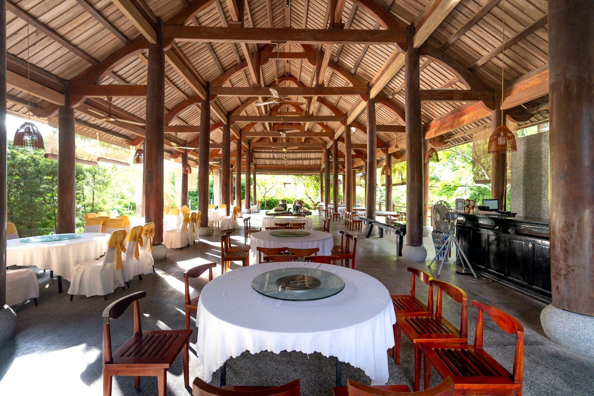 Open-air restaurant with round tables, white tablecloths, and wooden chairs under a wooden roof.