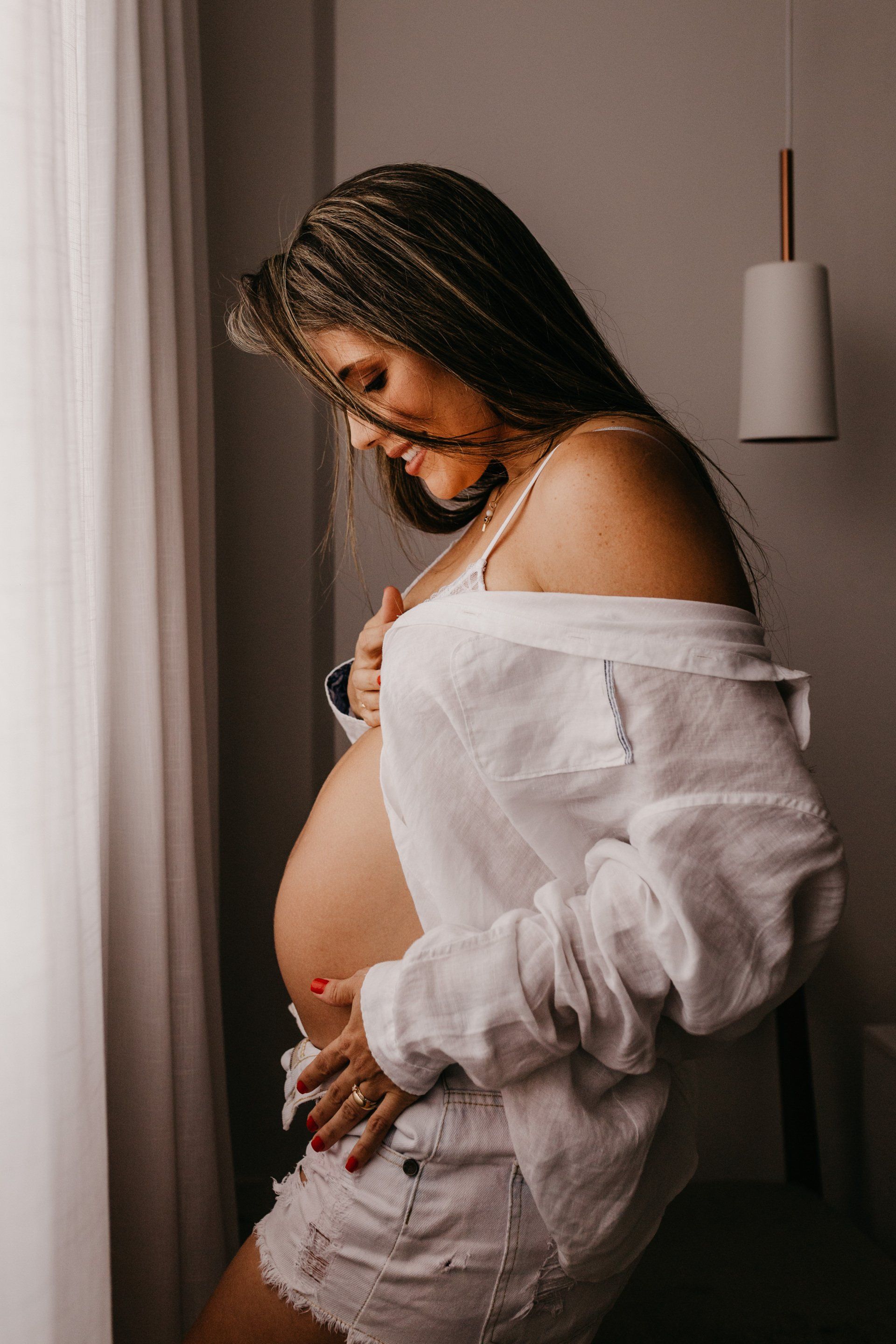 A pregnant woman is standing in front of a window holding her belly.