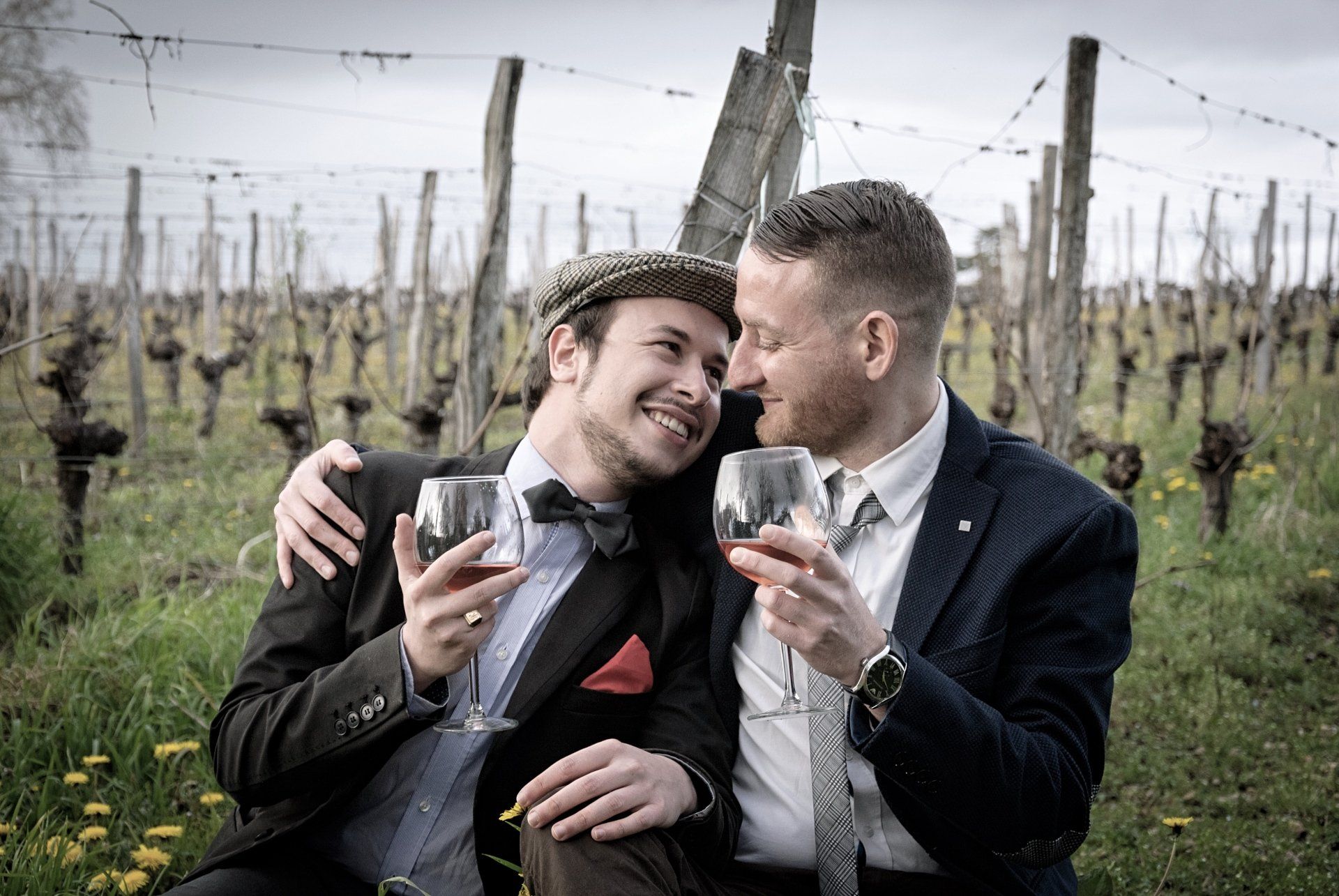 Two men are sitting next to each other in a vineyard holding wine glasses.