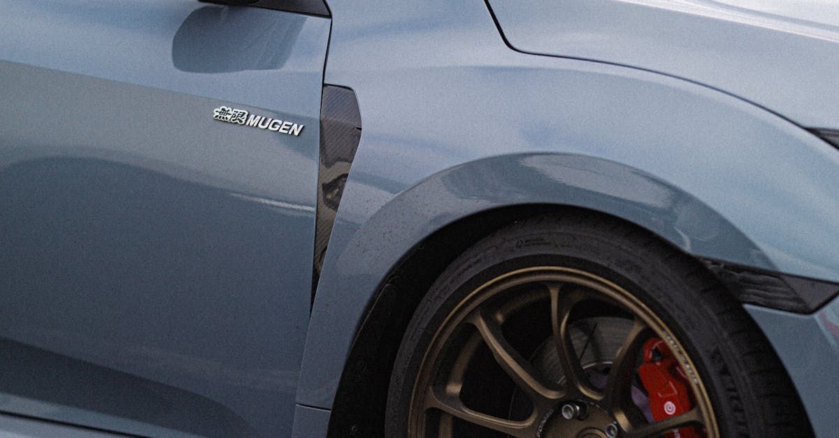 Close-up of a gray car's front wheel and fender, with a Mugen logo, bronze rim, and red brake caliper.