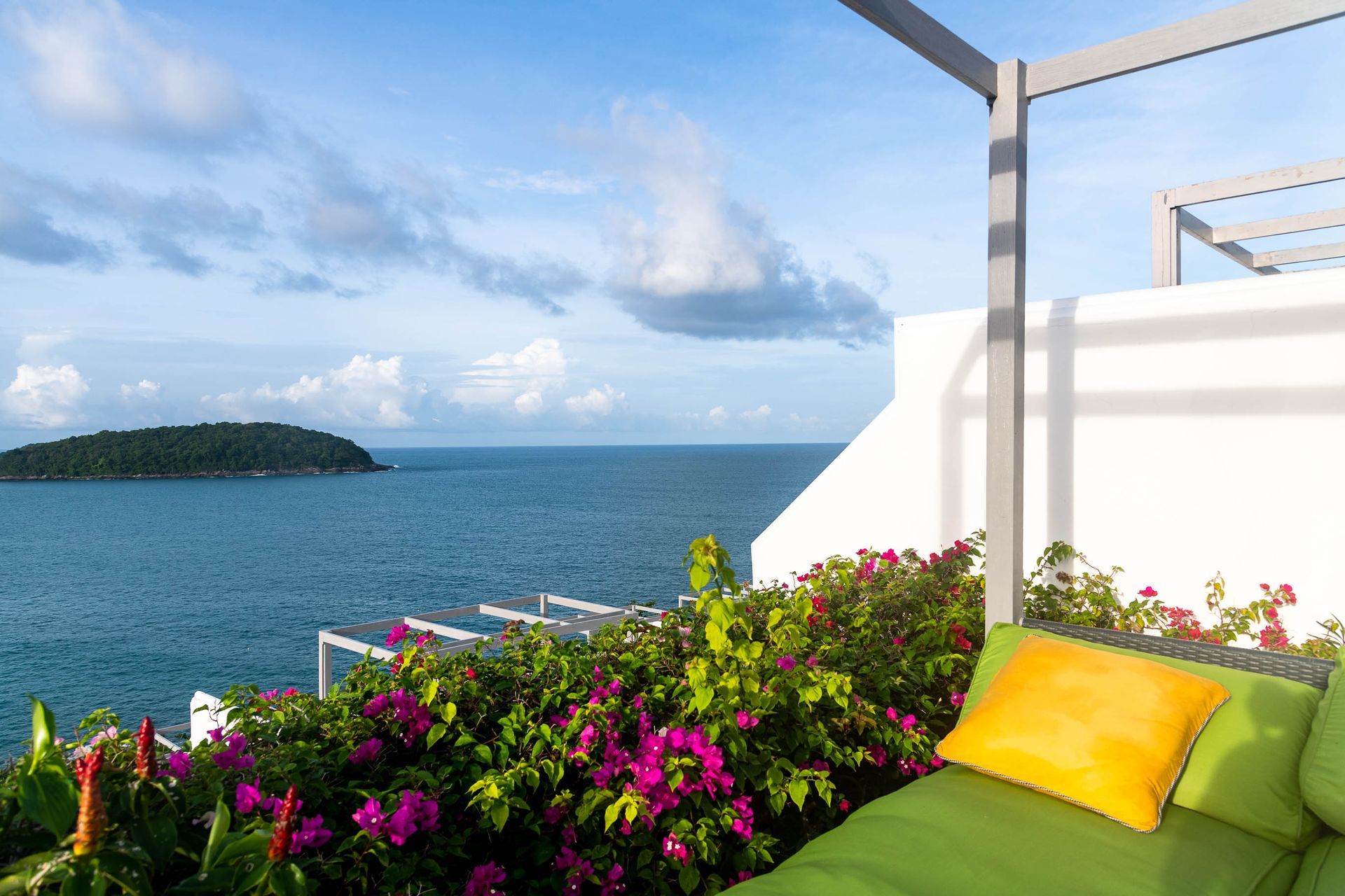 A green couch with a yellow pillow sits on a balcony overlooking the ocean.