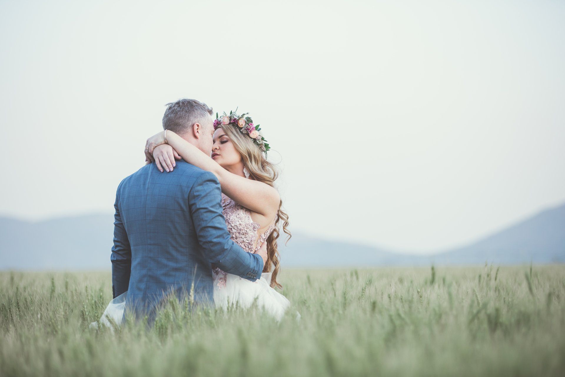 A bride and groom are sitting in a field of wheat and kissing.