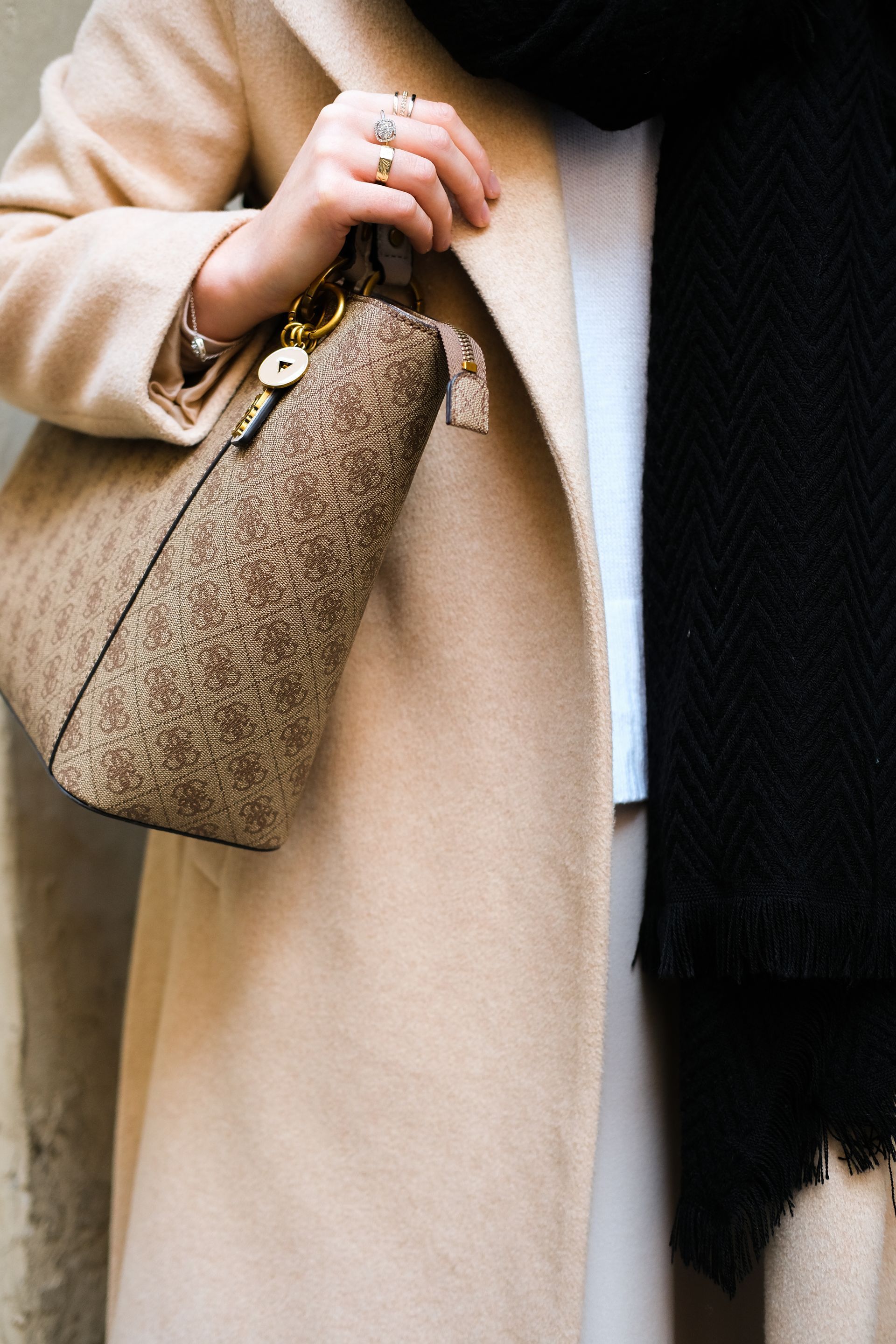 A woman wearing a tan coat and a black scarf is holding a purse.
