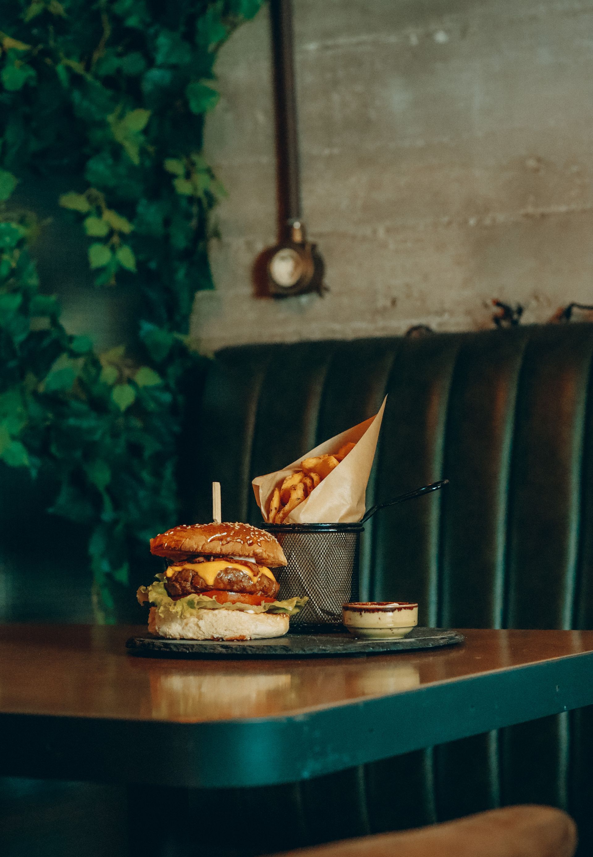 Burger with fries and dipping sauce on a table, against a green booth and wall with vines.