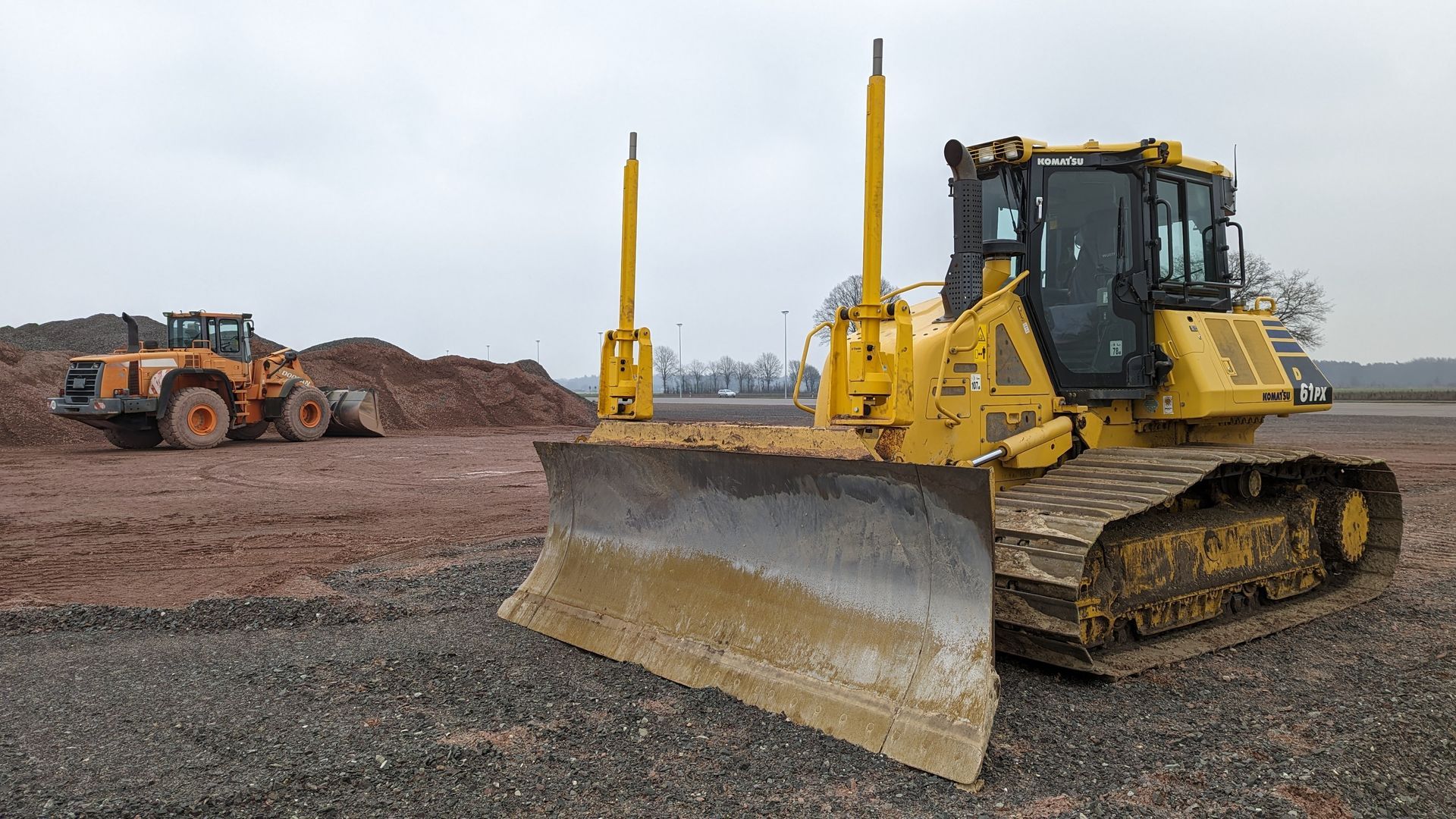 A bulldozer is parked in a gravel lot next to a tractor.