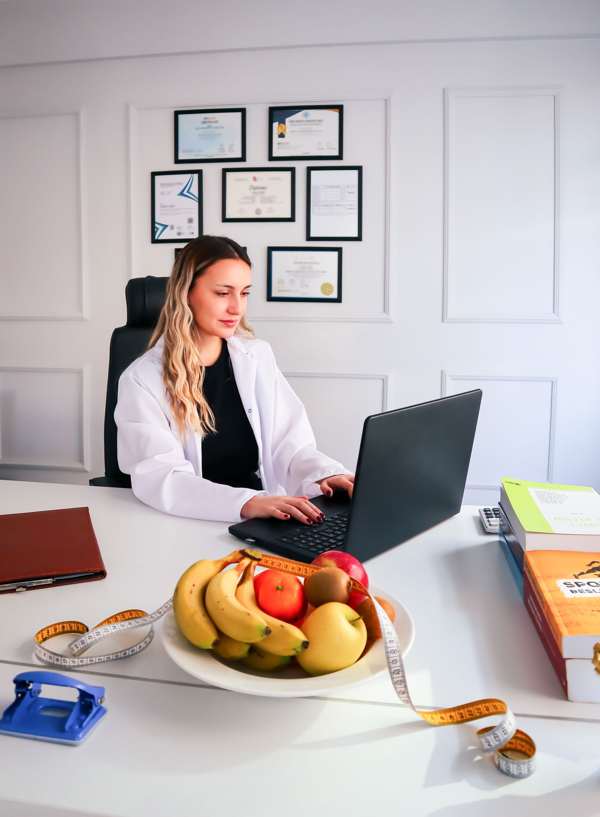 A woman is sitting at a desk with a laptop and a bowl of fruit.