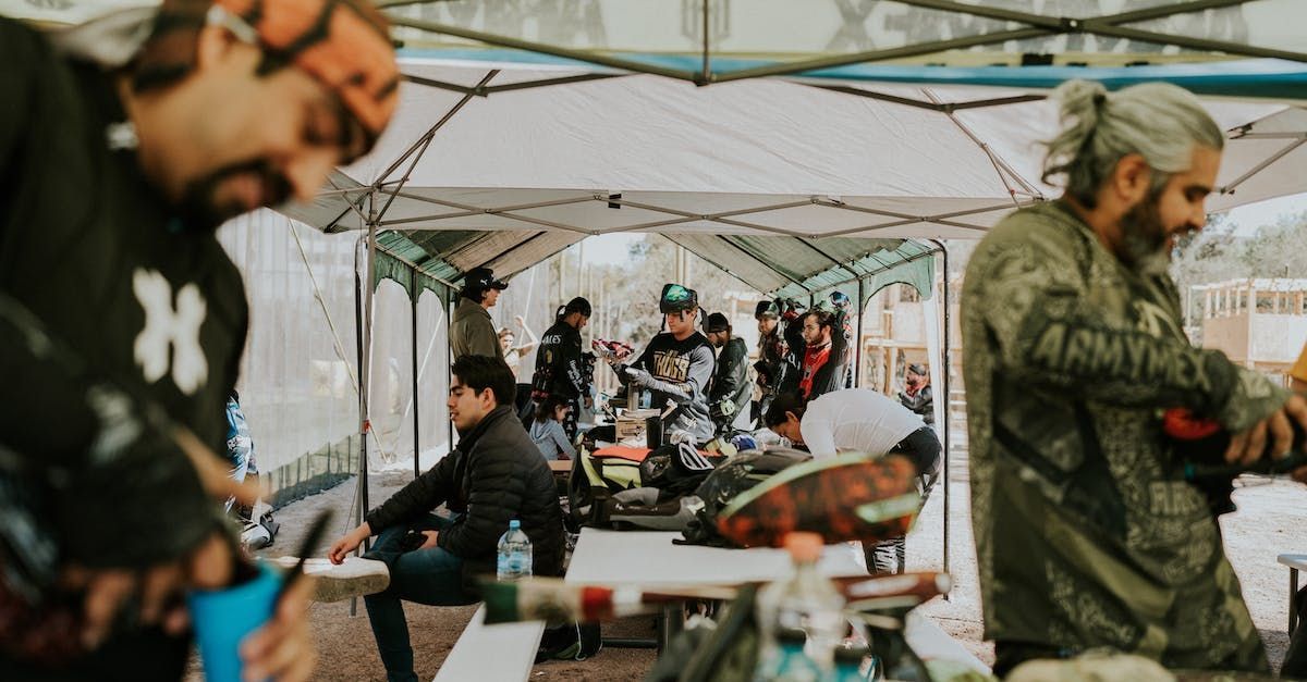 People at a paintball event under a canopy, preparing gear and resting.