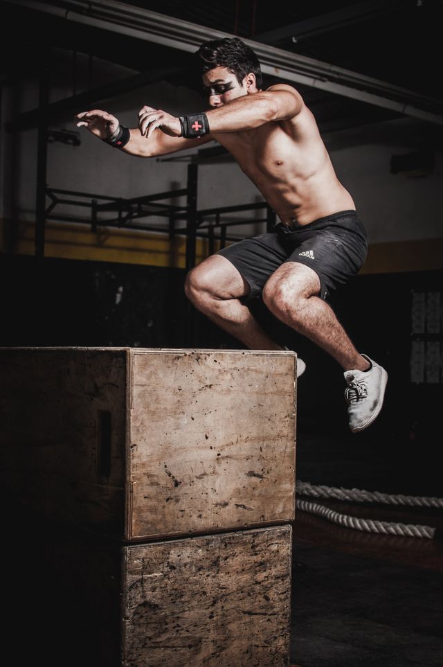 A shirtless man is jumping over a wooden box in a gym.