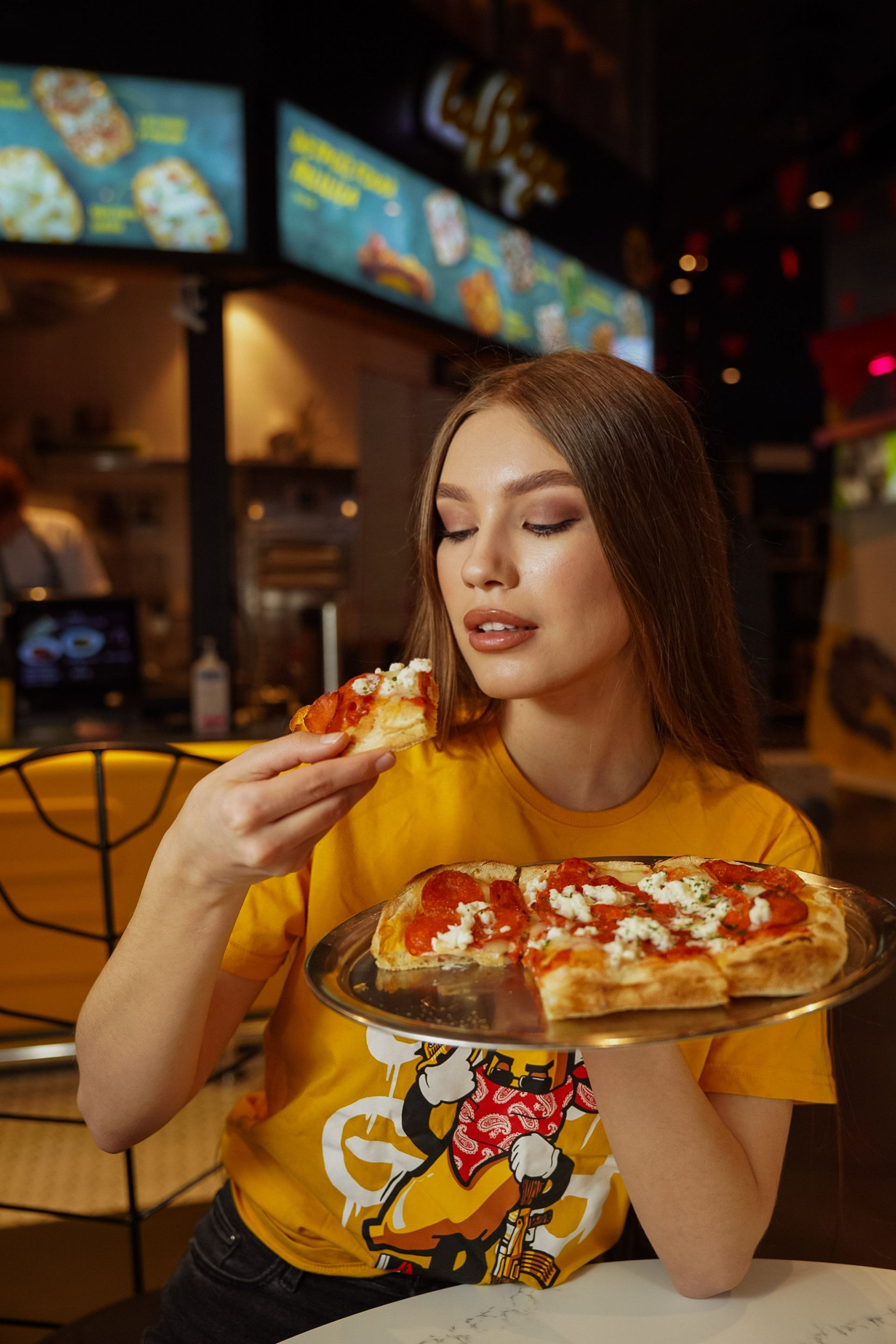 Woman eating pizza slice from a tray in a restaurant; yellow shirt, dark hair.