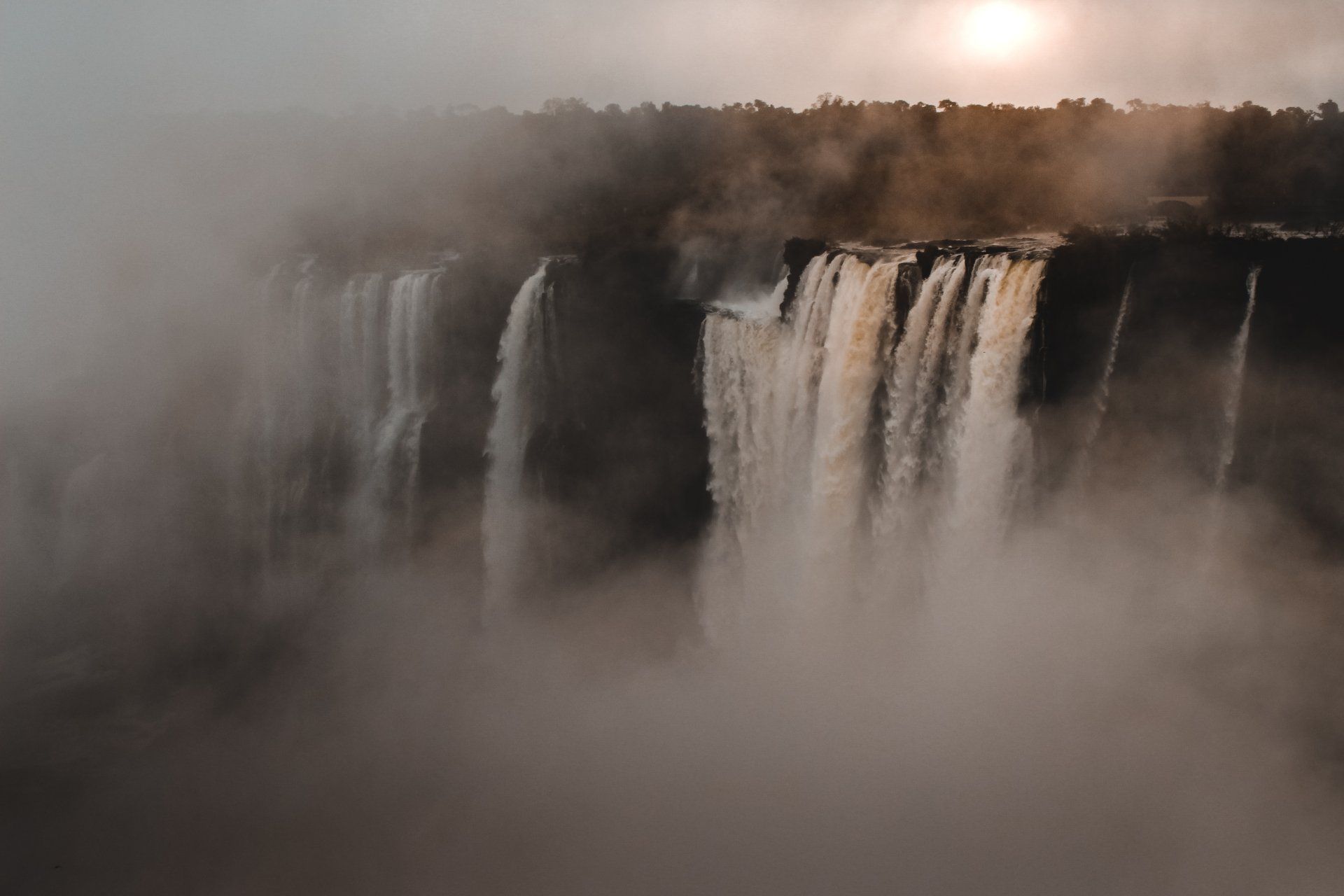 A waterfall is surrounded by fog and the sun is shining through the fog.