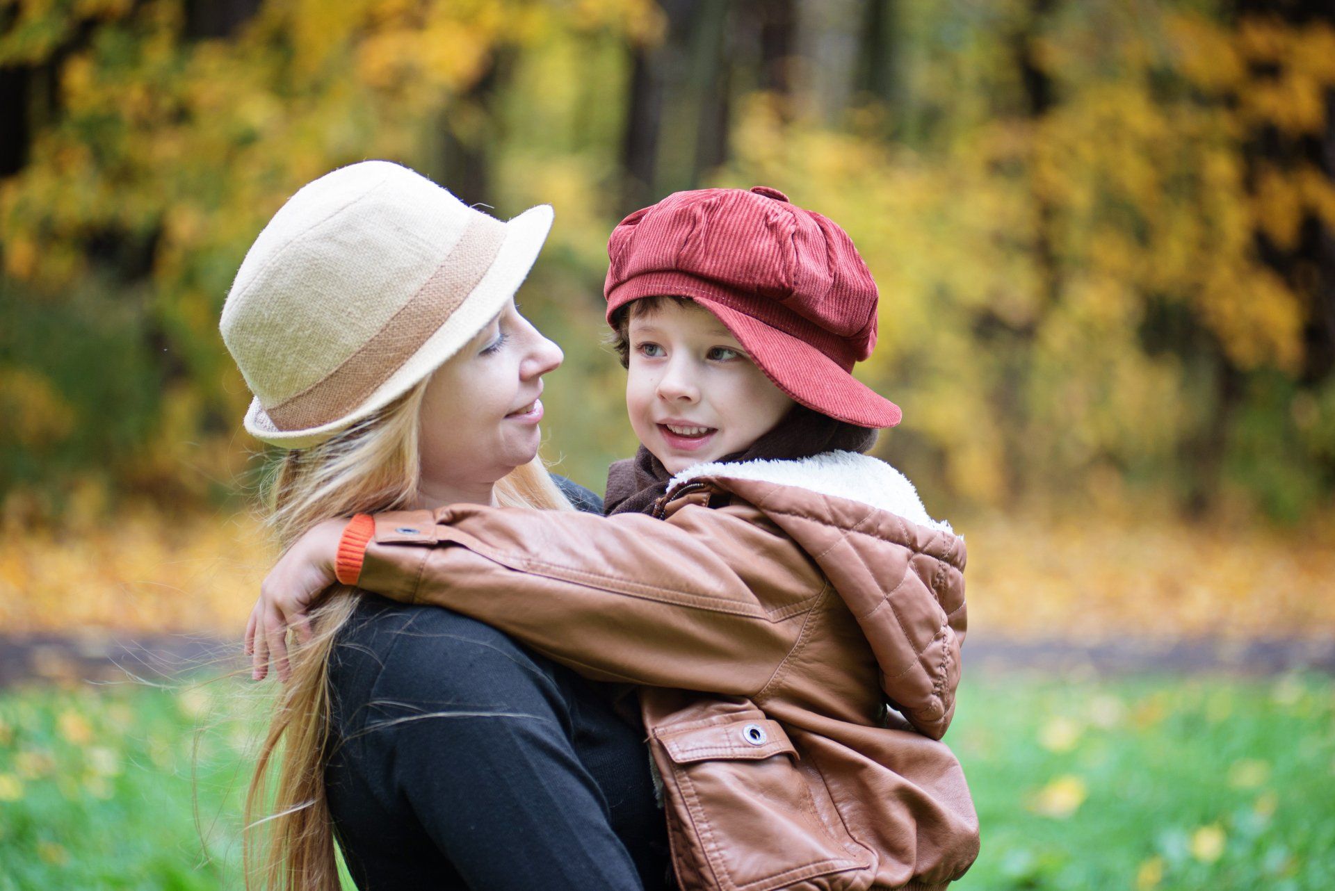 Woman with blonde hair hugs a child wearing a red hat outdoors in autumn.