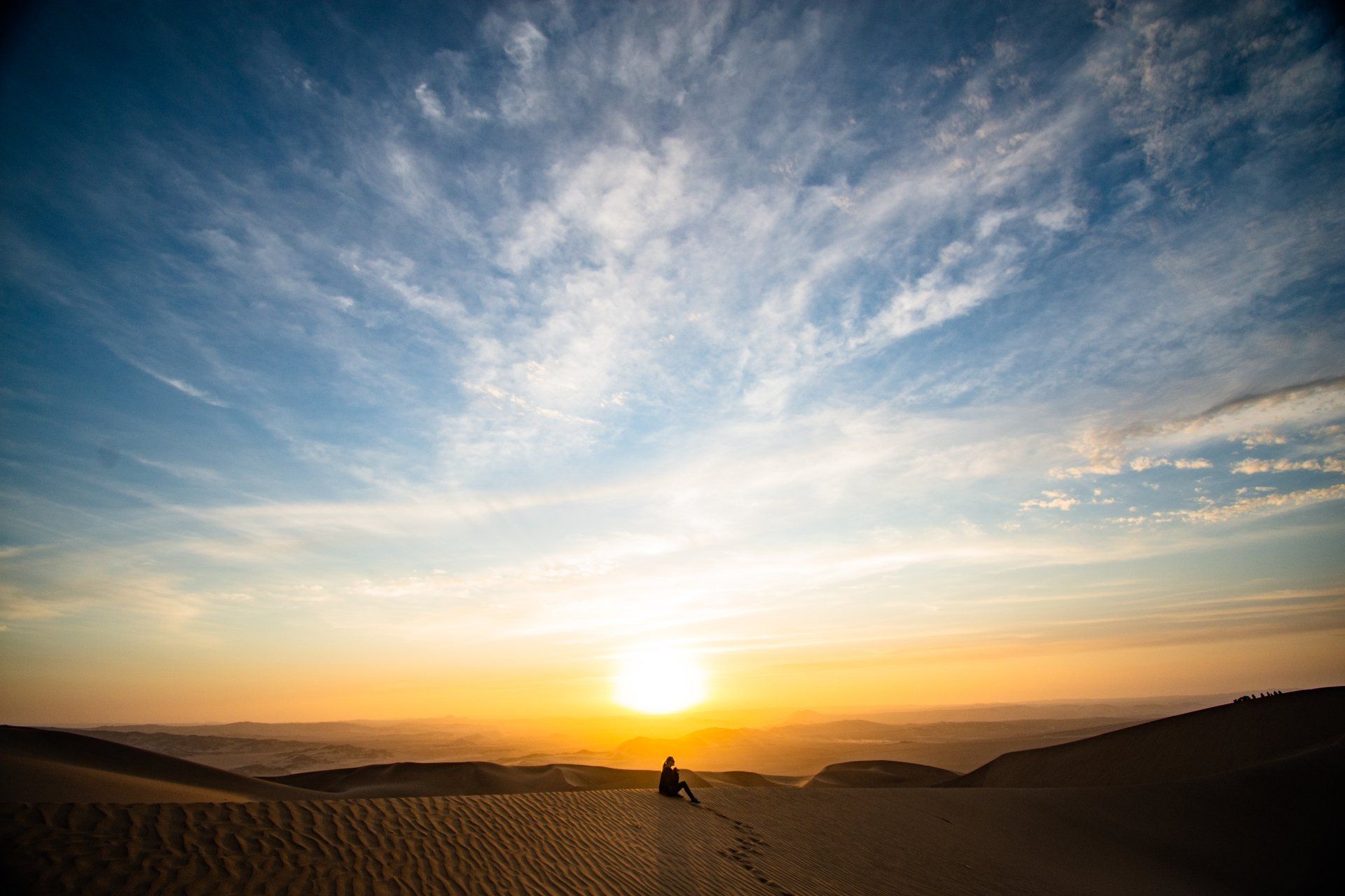 A person is sitting on top of a sand dune at sunset.