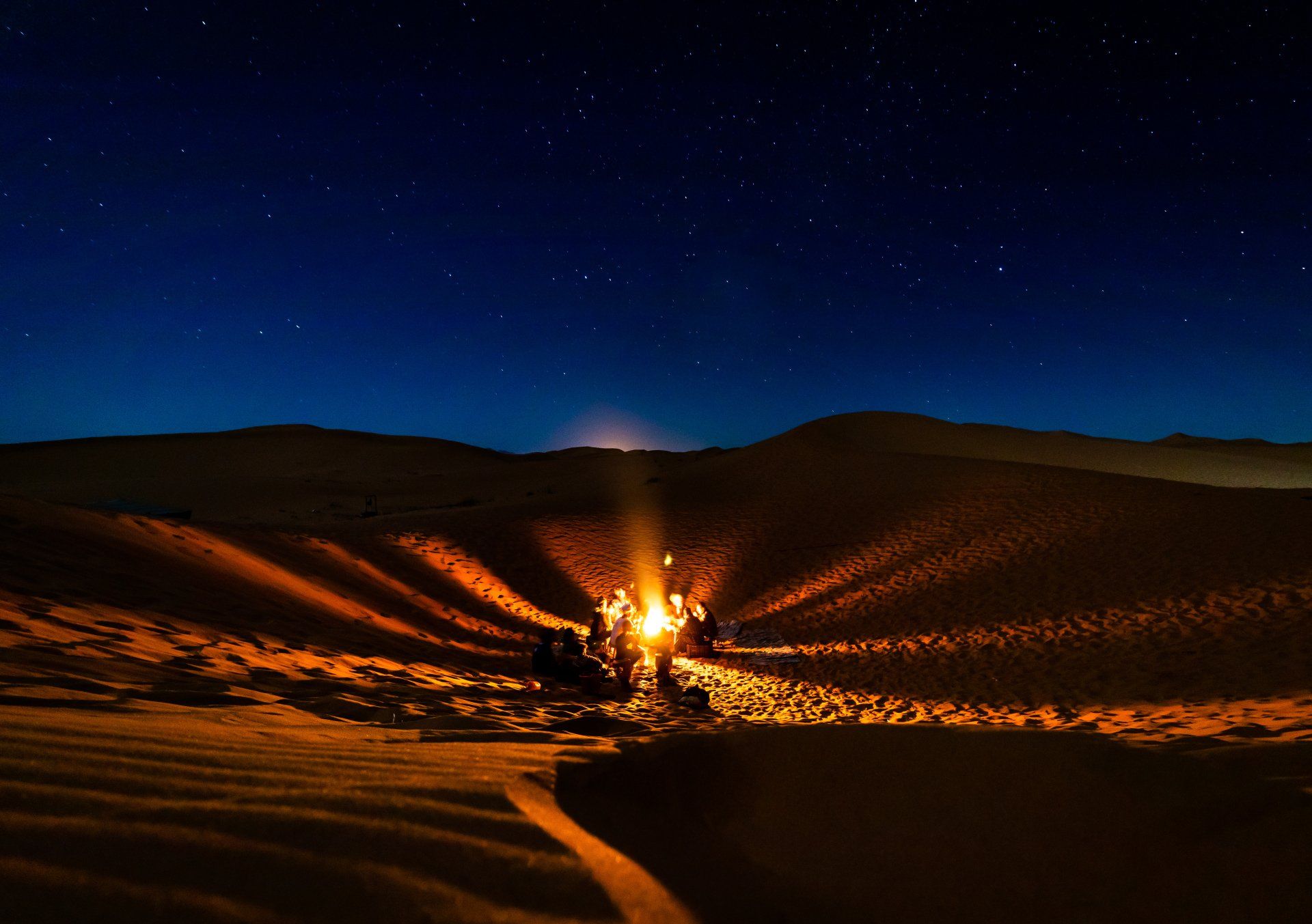 A group of people are sitting around a campfire in the middle of a desert at night.