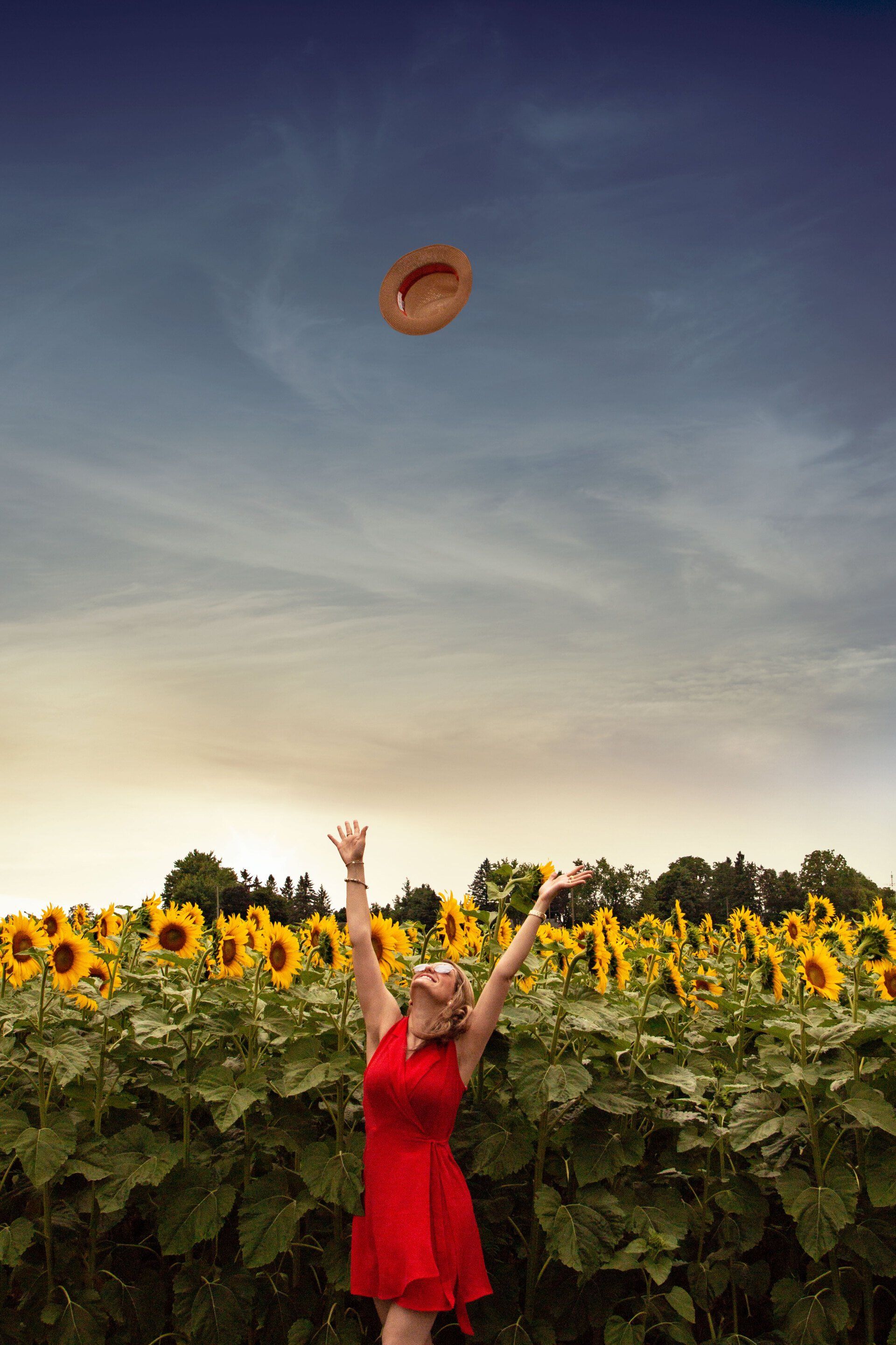 Een vrouw in een rode jurk gooit een hoed in de lucht in een veld met zonnebloemen.