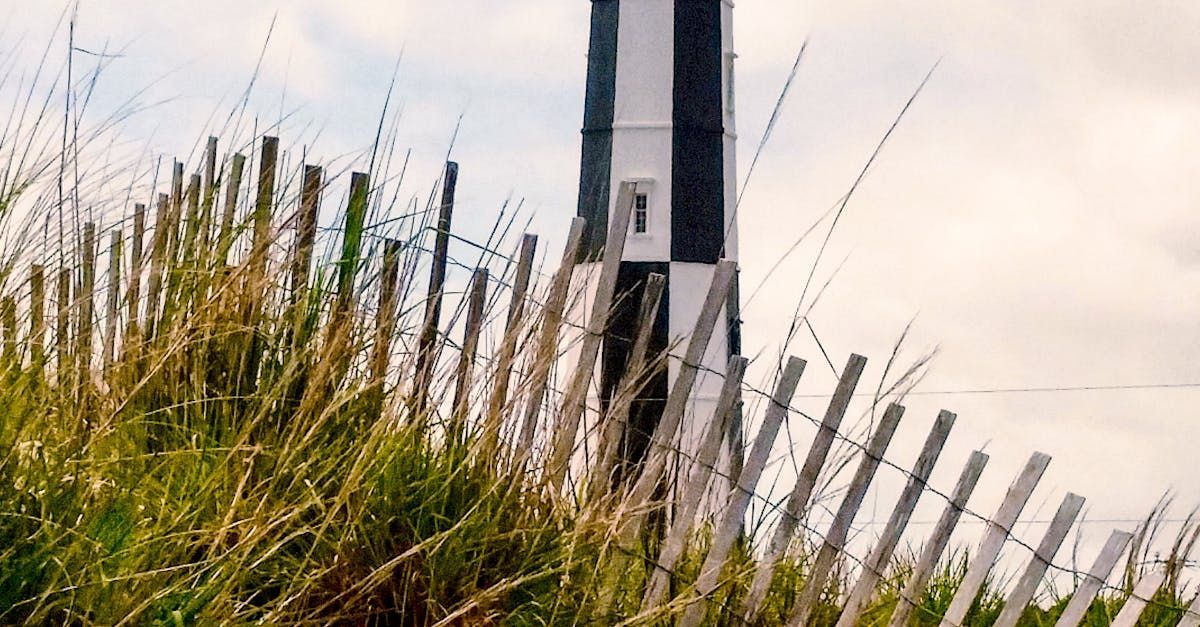 A lighthouse is behind a wooden fence on top of a grassy hill.