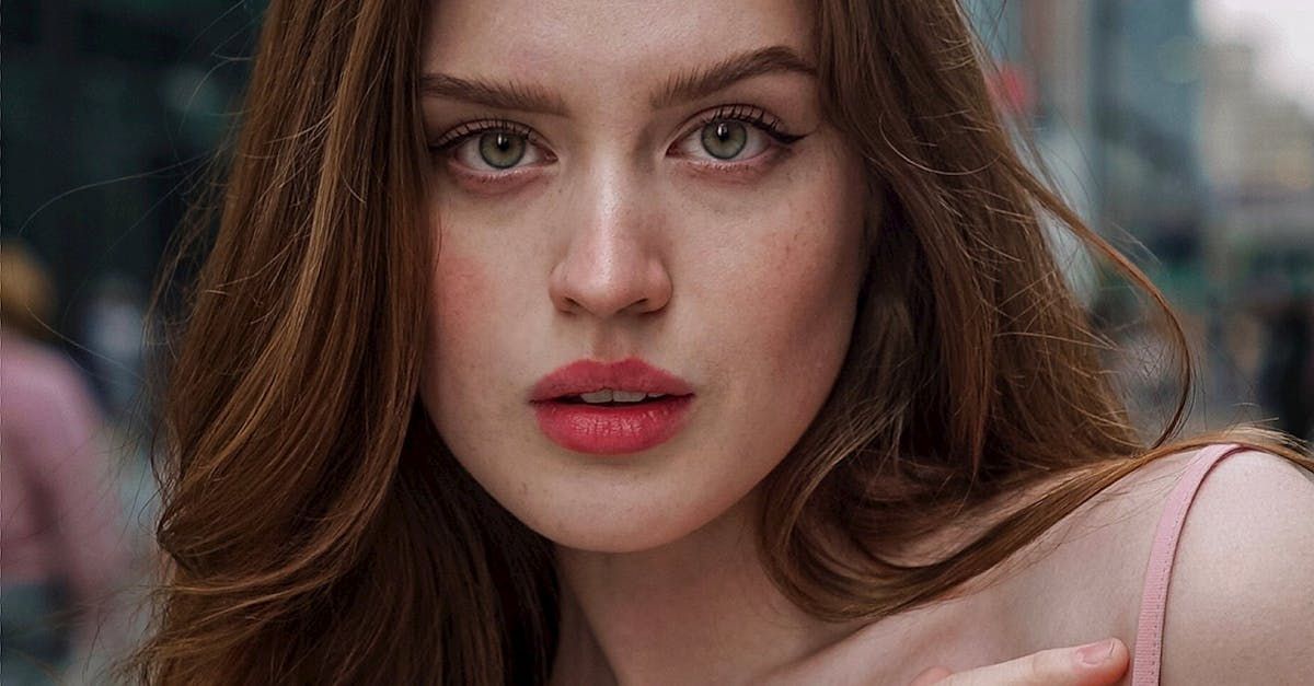 Young woman with auburn hair, pale skin, pink top, and subtle makeup, looking directly at the camera in an urban setting.