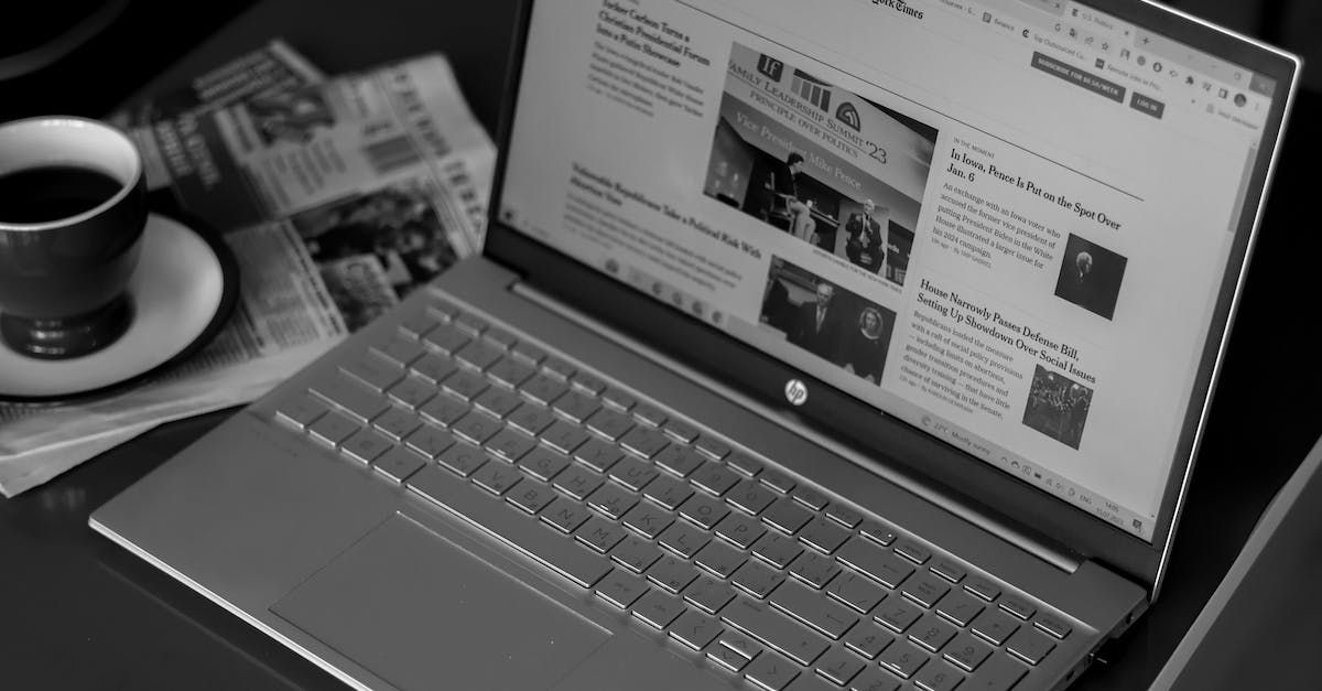 A laptop computer is open on a table next to a cup of coffee and a newspaper.