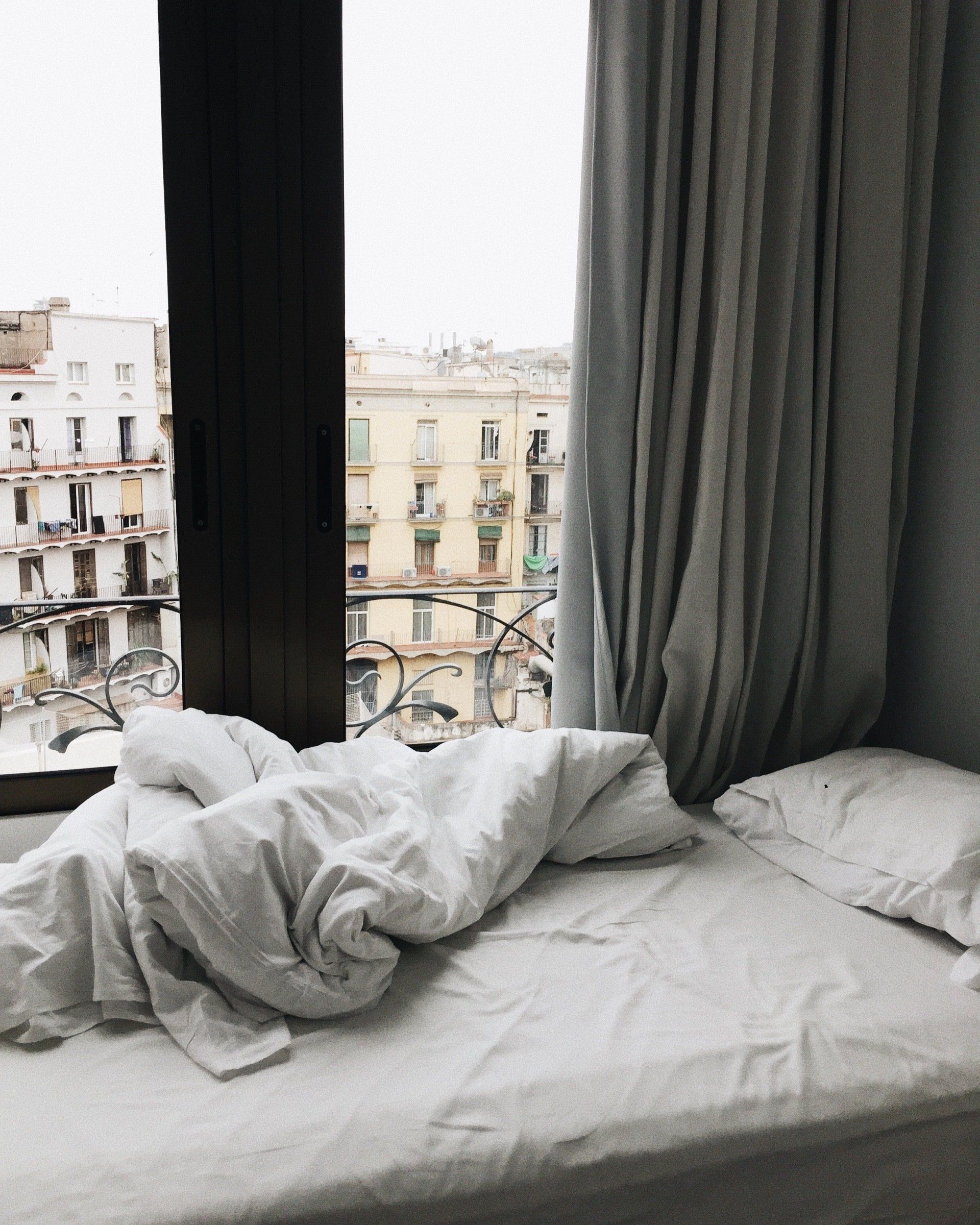 Unmade bed by a window overlooking a city. White sheets and blanket, gray curtains.