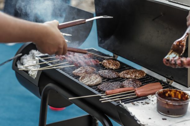 Hands grilling assorted meat skewers and patties over a smoky barbecue grill