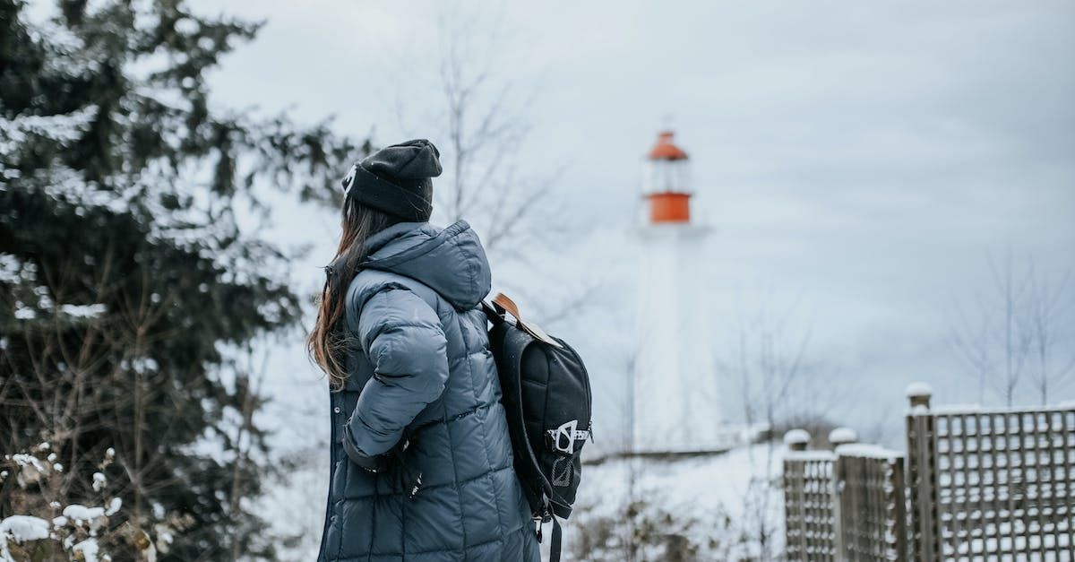 A person in a winter coat and backpack standing in front of a lighthouse in the snow.