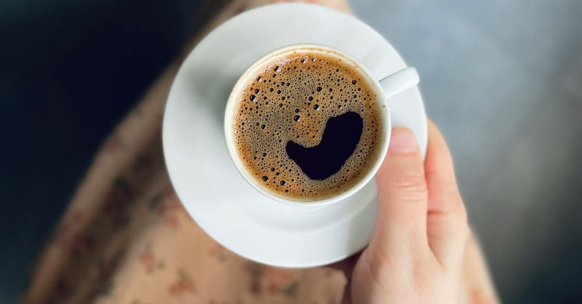 Hand holding a white teacup of coffee with a heart-shaped crema on a saucer, against a blurred background.