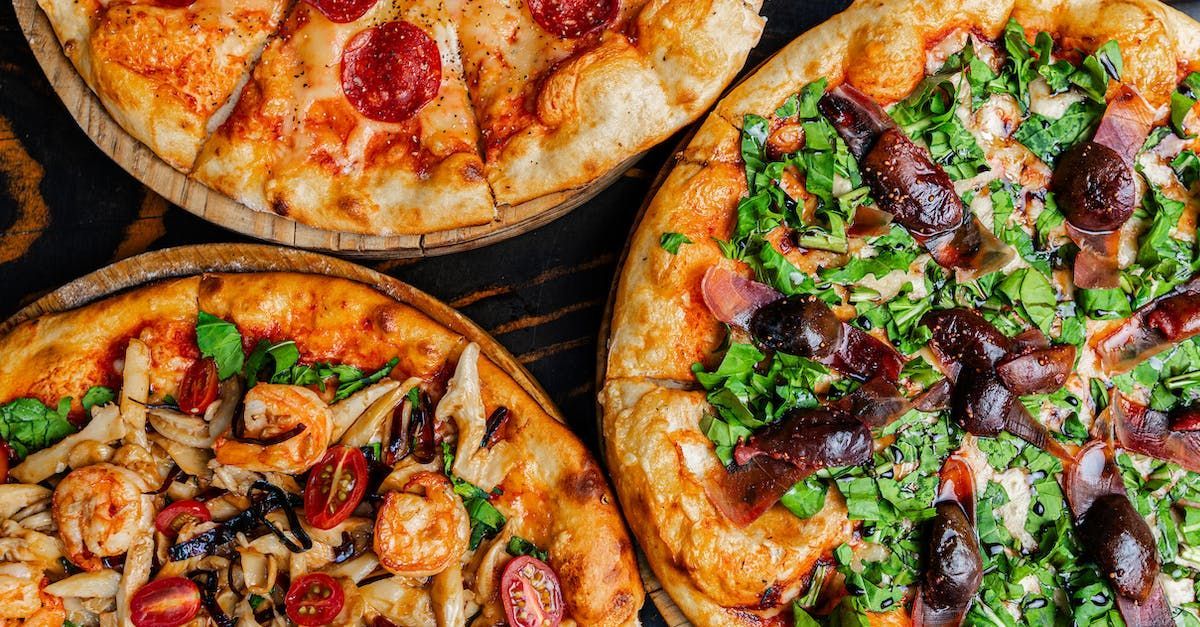 Tre pizze: pepperoni, datteri e verdure, gamberi e funghi, su un tavolo di legno scuro.