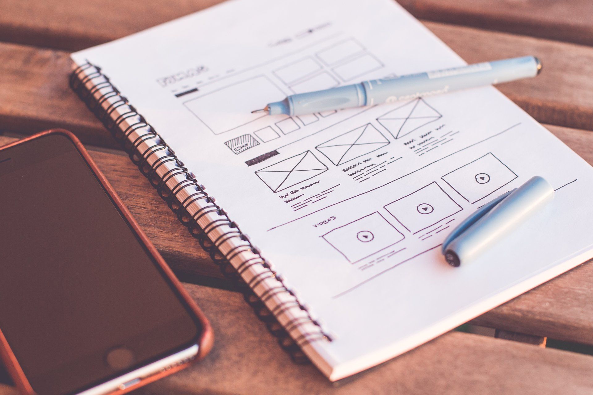 A notebook with pens and a cell phone on a wooden table.