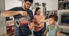 A man is pouring a smoothie into a glass in a kitchen while a woman holds a baby.